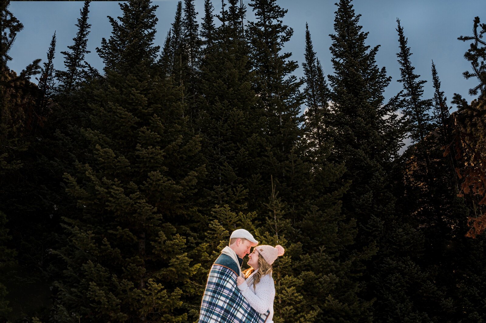 Couple wrapped up in a blanket and surrounded by pine trees during their winter engagement session.
