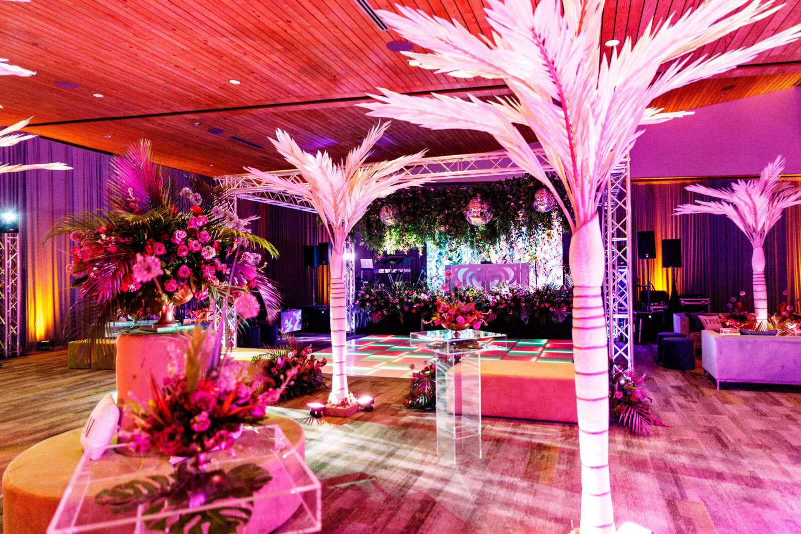 Tropical-themed event lounge with vibrant pink and purple floral arrangements, illuminated palm trees, LED dance floor, disco balls, and lush greenery backdrop—designed by Wells Design Co for a luxury celebration.