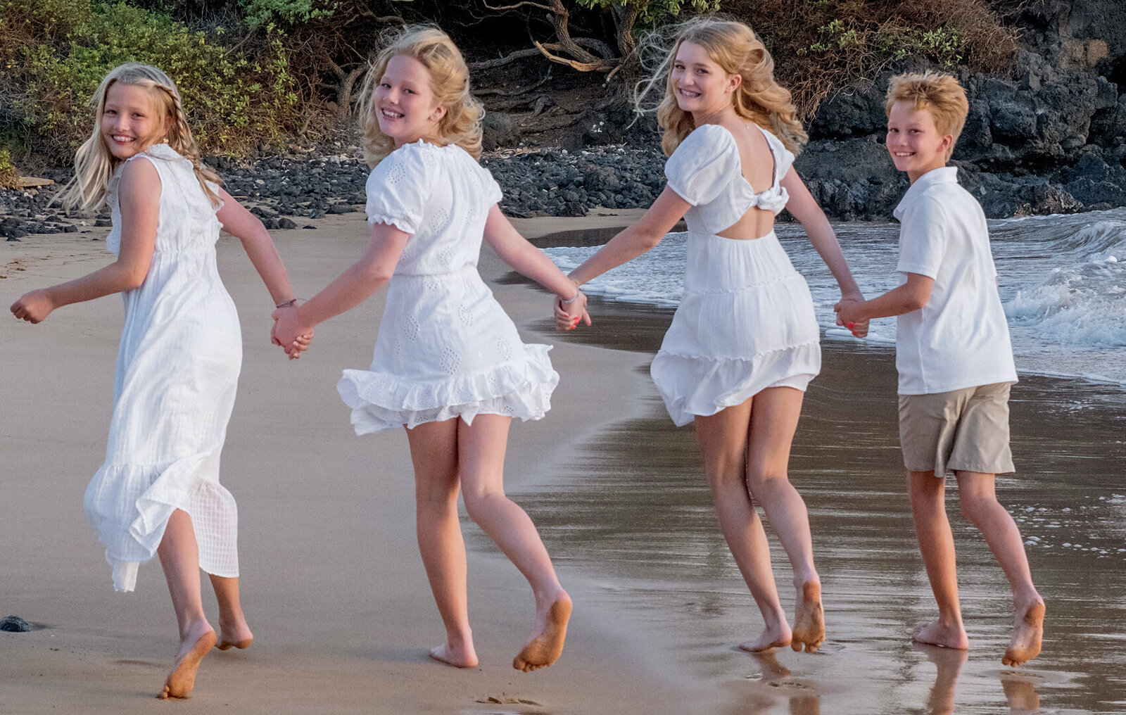 children, family of four, three sisters and one brother all wearing white and running along the beach in Oahu, looking over their shoulder back at the camera. 
