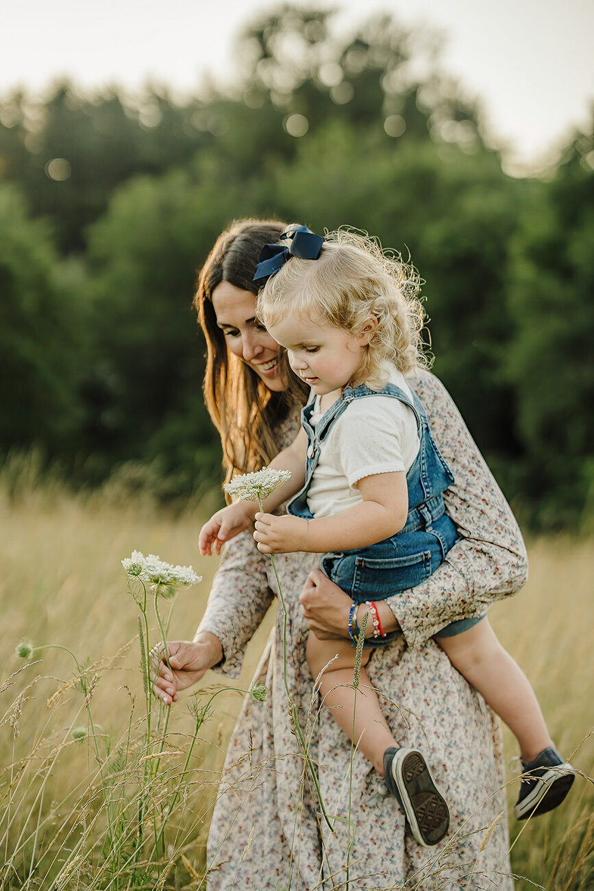 mom holds toddler daughter in jean overalls while picking flowers