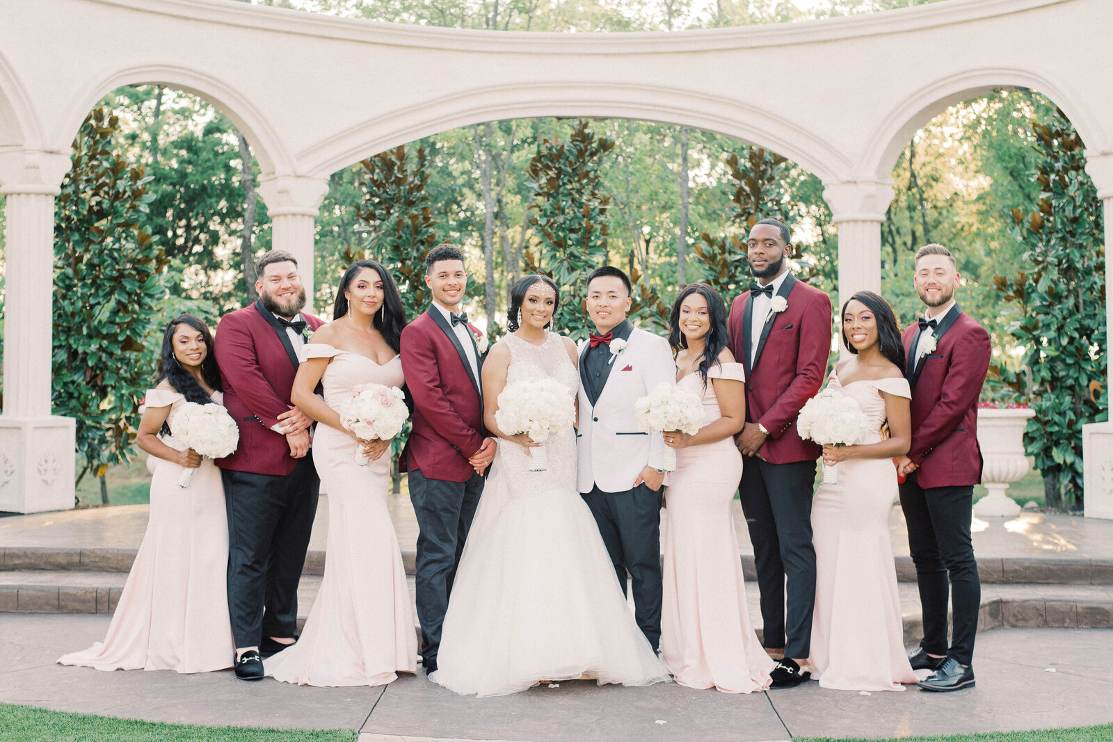 Bride and groom smiling and standing with the wedding party bridesmaids in light pink dresses and groomsmen in deep red suits