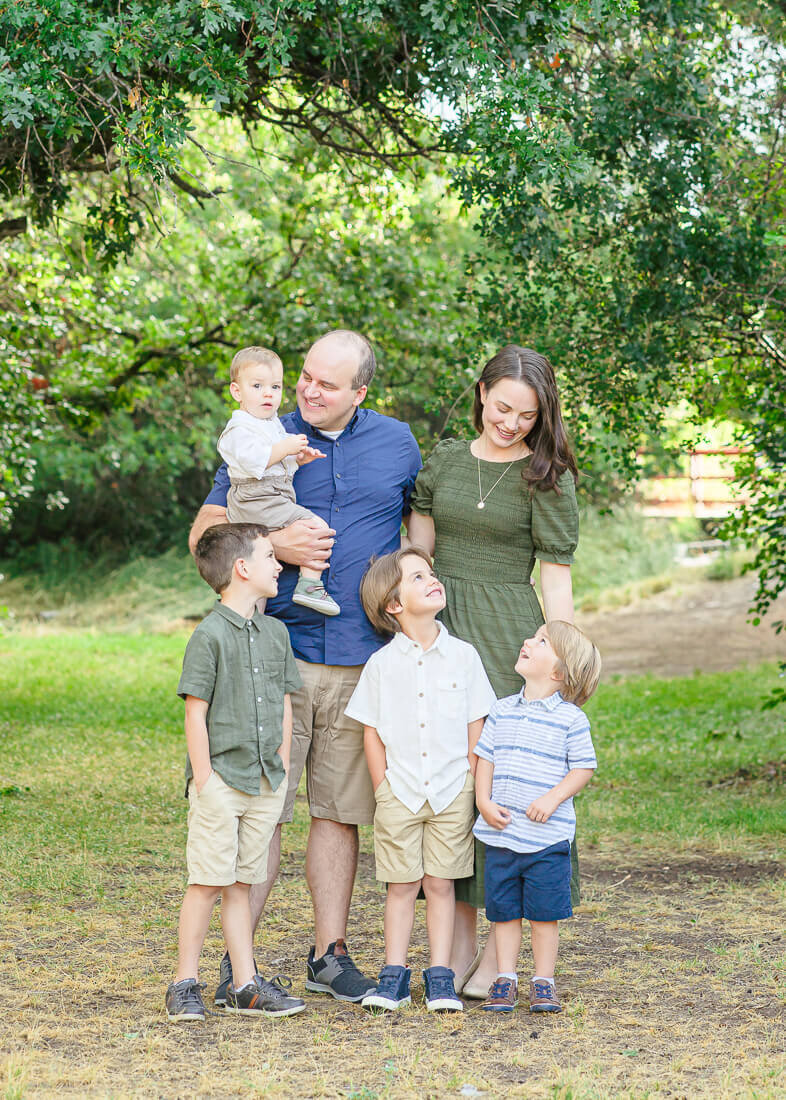 Salt Lake Photography of a father, mother, and their four young boys look at each other smiling while standing in front of lush green oak trees in Highland Glen Park, Lehi.