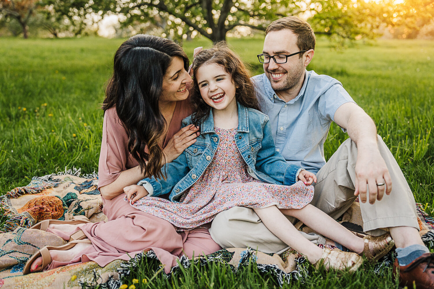 mom and dad laugh with daughter in a field of blossoms at sunset