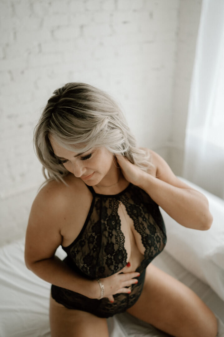 Blonde woman kneeling on white bed looking down, wearing black lace lingerie