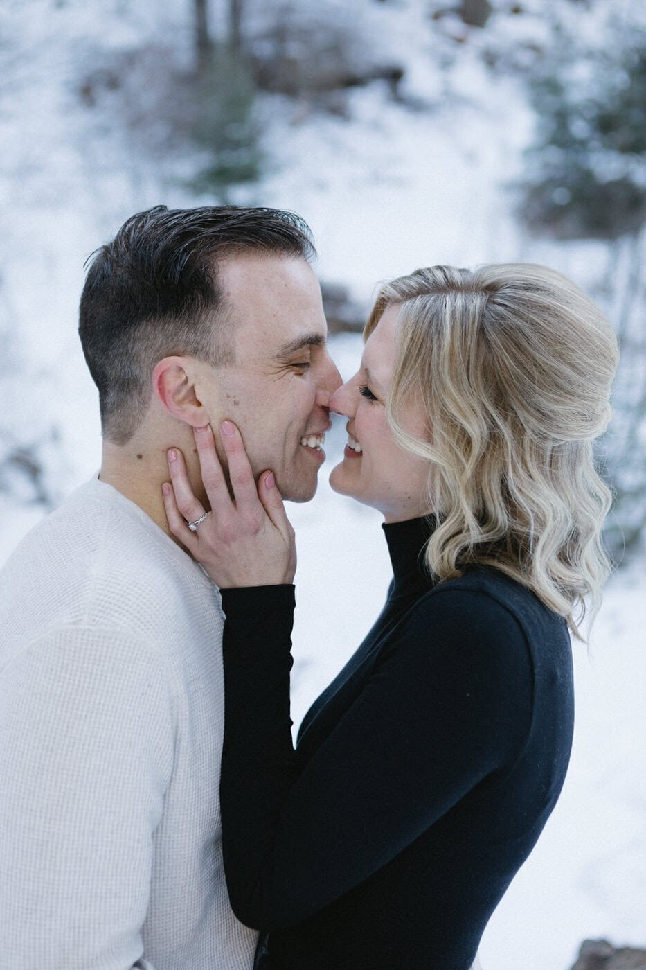 A couple stands nose to nose smiling at each other with snow in the background.
