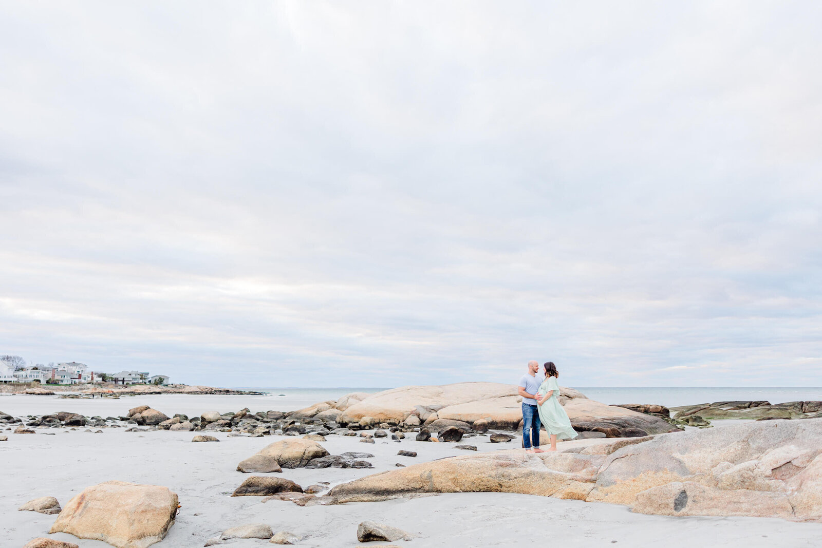 Epic view of a pregnant woman and her husband sharing a moment on top of a large rock with the ocean and cloudy sky in the background