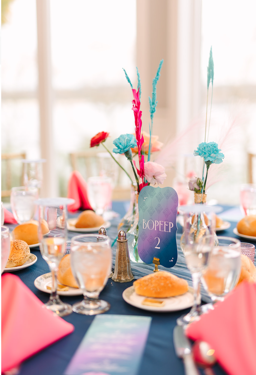 A navy wedding reception table with hot pink napkins and rainbow wedding manus.