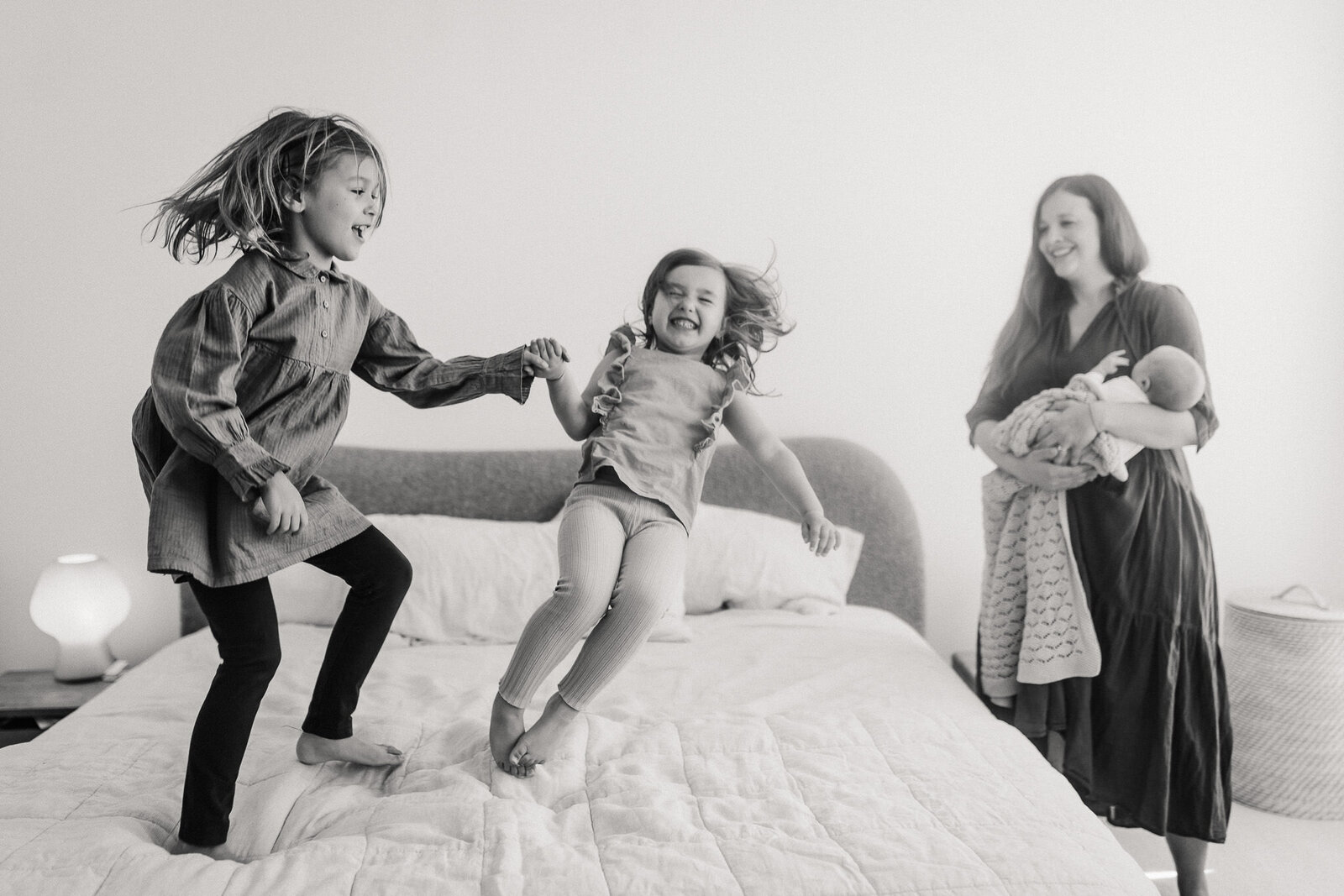 in home lifestyle newborn session with mom holding baby and sisters jumping on bed