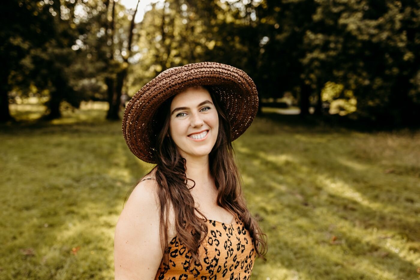 Smiling woman with long brown hair and a wide-brimmed straw hat, wearing a leopard-print top, outdoors on a sunny day.