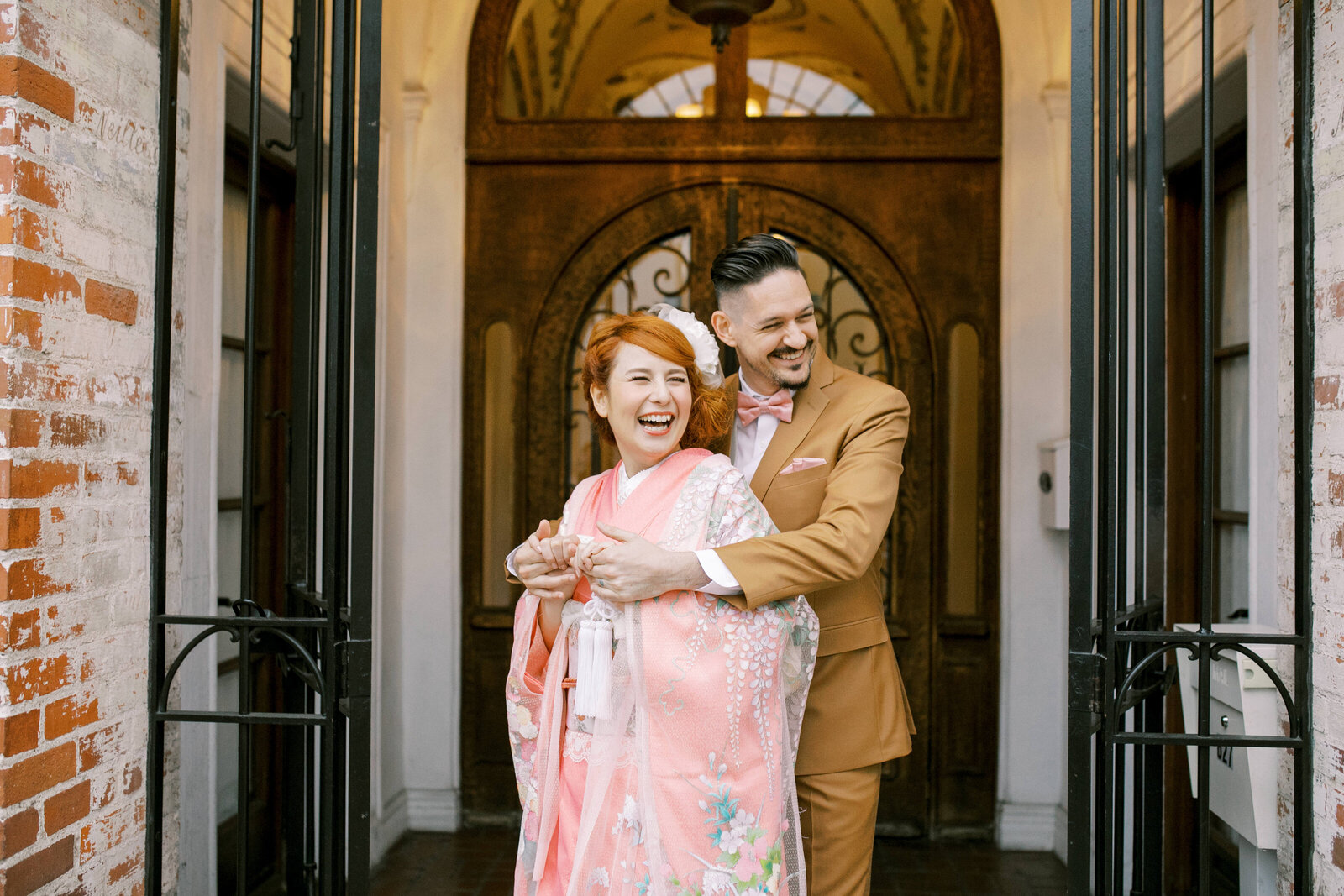 Laughing couple at Carondelet House, bride in soft pink irouchikake and groom embracing by archway, Los Angeles California. Lovely wedding kimono rental shoot | カロンデレットハウスのアーチのもとで微笑む淡いピンク振袖花嫁とキャメルスーツの新郎。ロサンゼルス カリフォルニアの着物レンタル。
