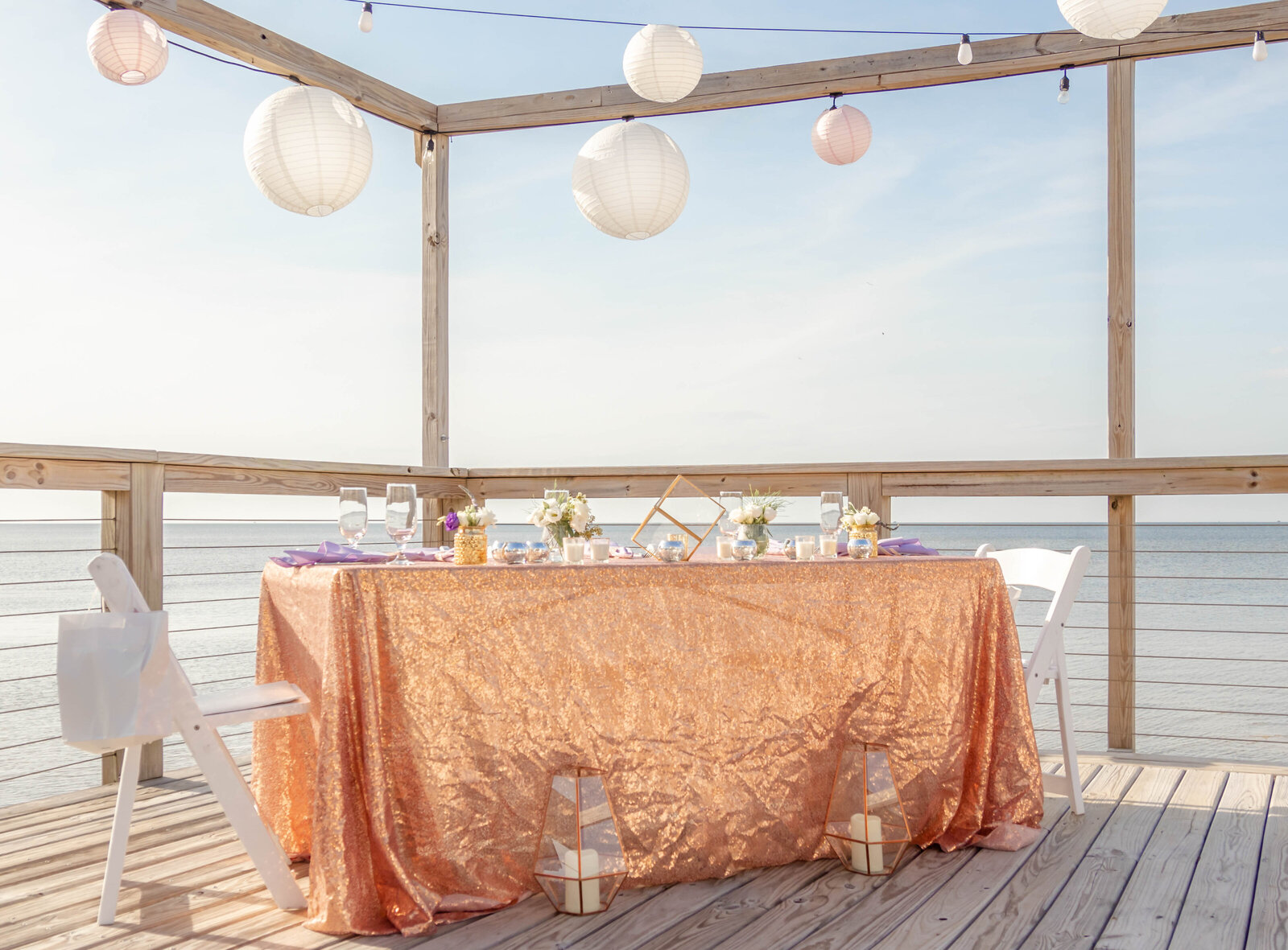 A beautifully decorated wedding table with a sparkling rose gold tablecloth, floral centerpieces, and hanging lanterns, set on a waterfront deck in Hatteras Island in the beautilf Outer Banks of North Carolina, creating a rustic yet elegant ambiance.