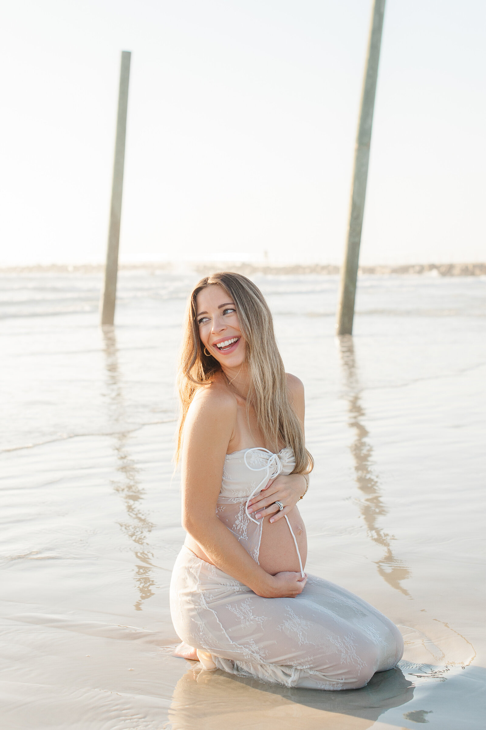 Pregnant mother holding her belly and sitting in the waves laughing over her shoulder