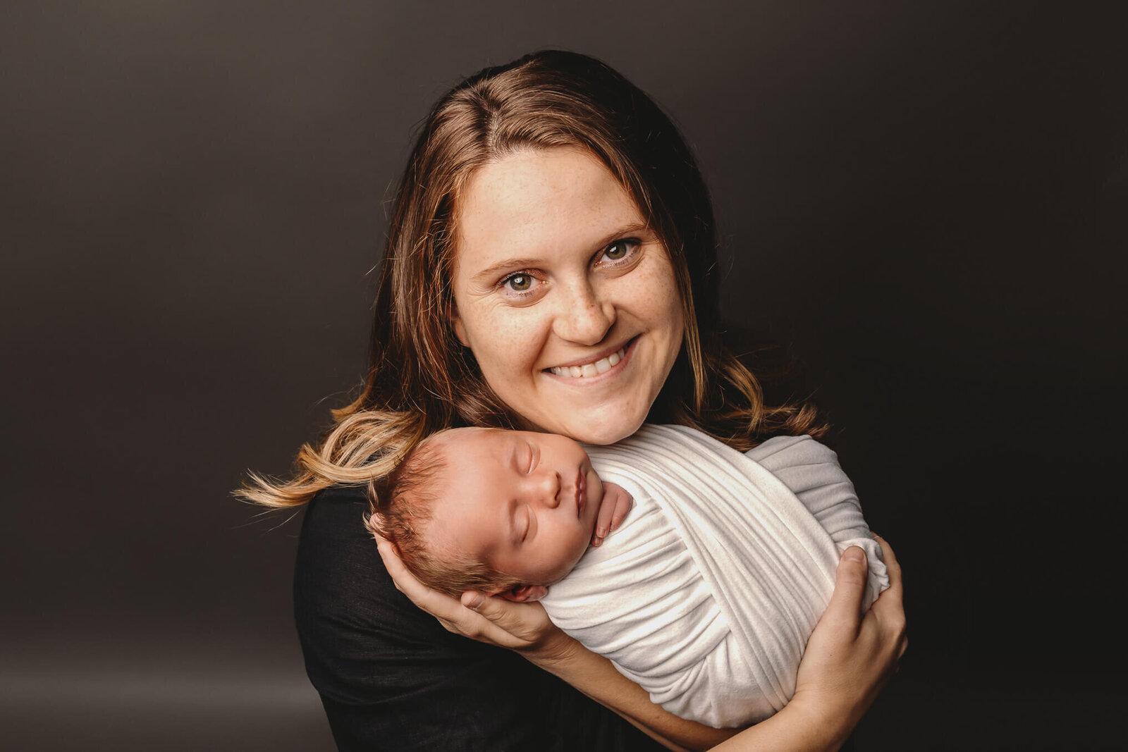 Mom in a black shirt smiling holding swaddled newborn baby in studio