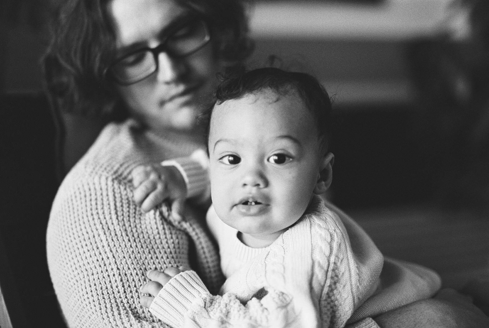 A tender black-and-white close-up of a mother holding her baby, with the child gazing curiously into the camera, radiating warmth and intimacy.