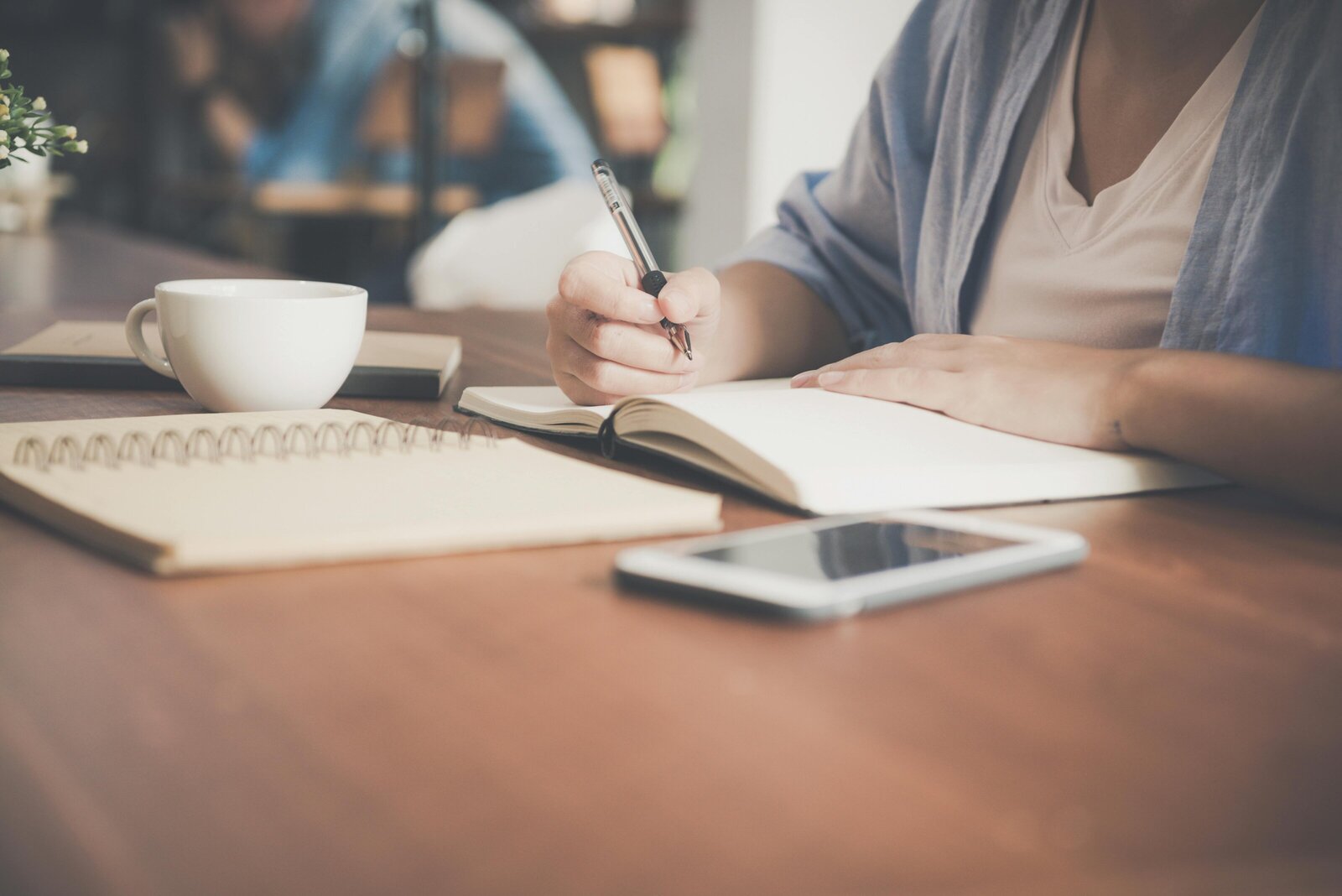 Person writing in a notebook at a desk with a smartphone and coffee nearby, suggesting financial planning or consultation session