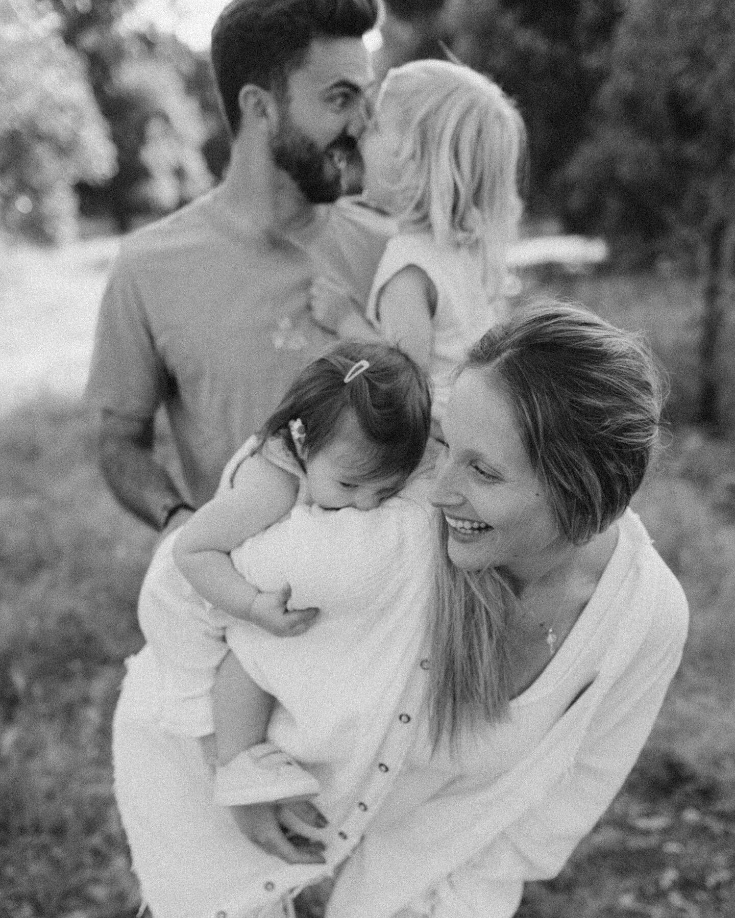 A black and white image of a family outdoors: a smiling woman holding a baby while a man, looking at a little girl on his shoulders, appears joyful.