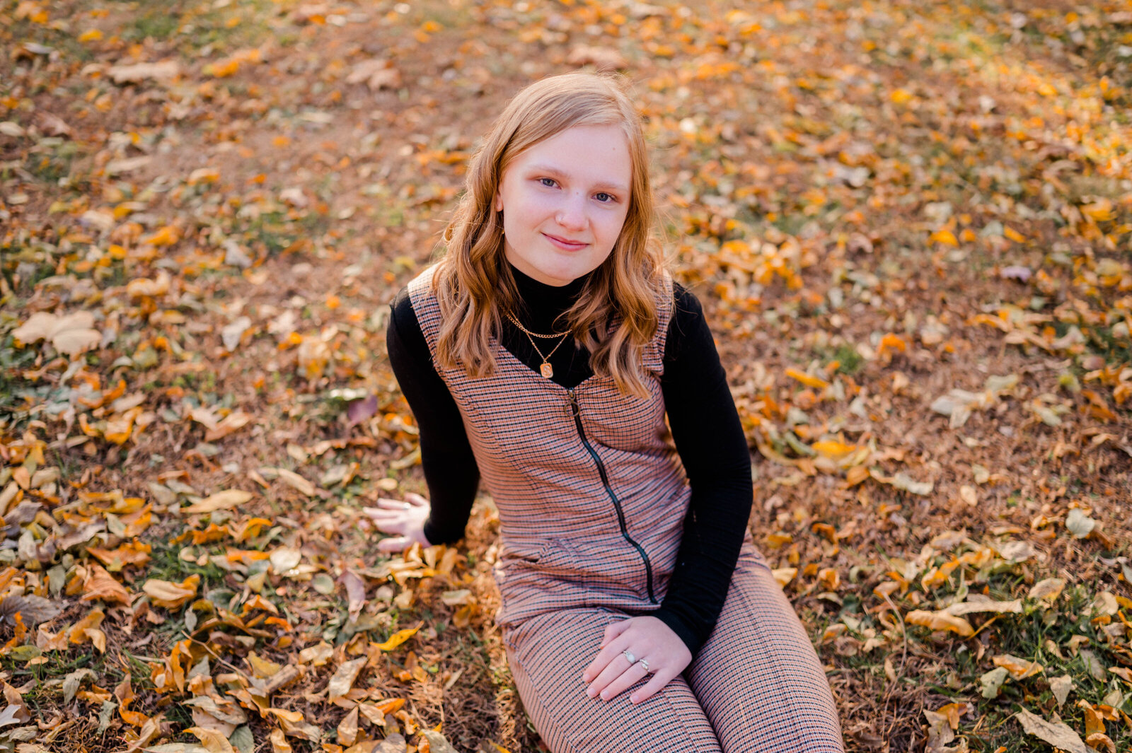 Teenage girl poses for the camera sitting on fallen autumn leaves.