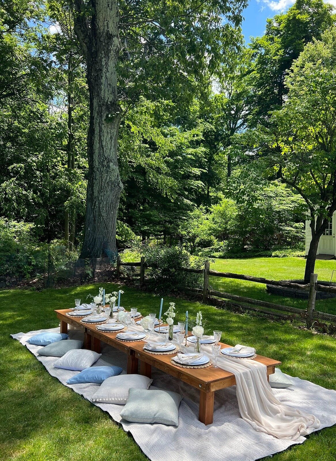 Backyard picnic table styled with blue and white tableware, candles, and cozy cushions in a shaded garden, designed by Yours Truly, a wedding planner and designer in New Jersey.