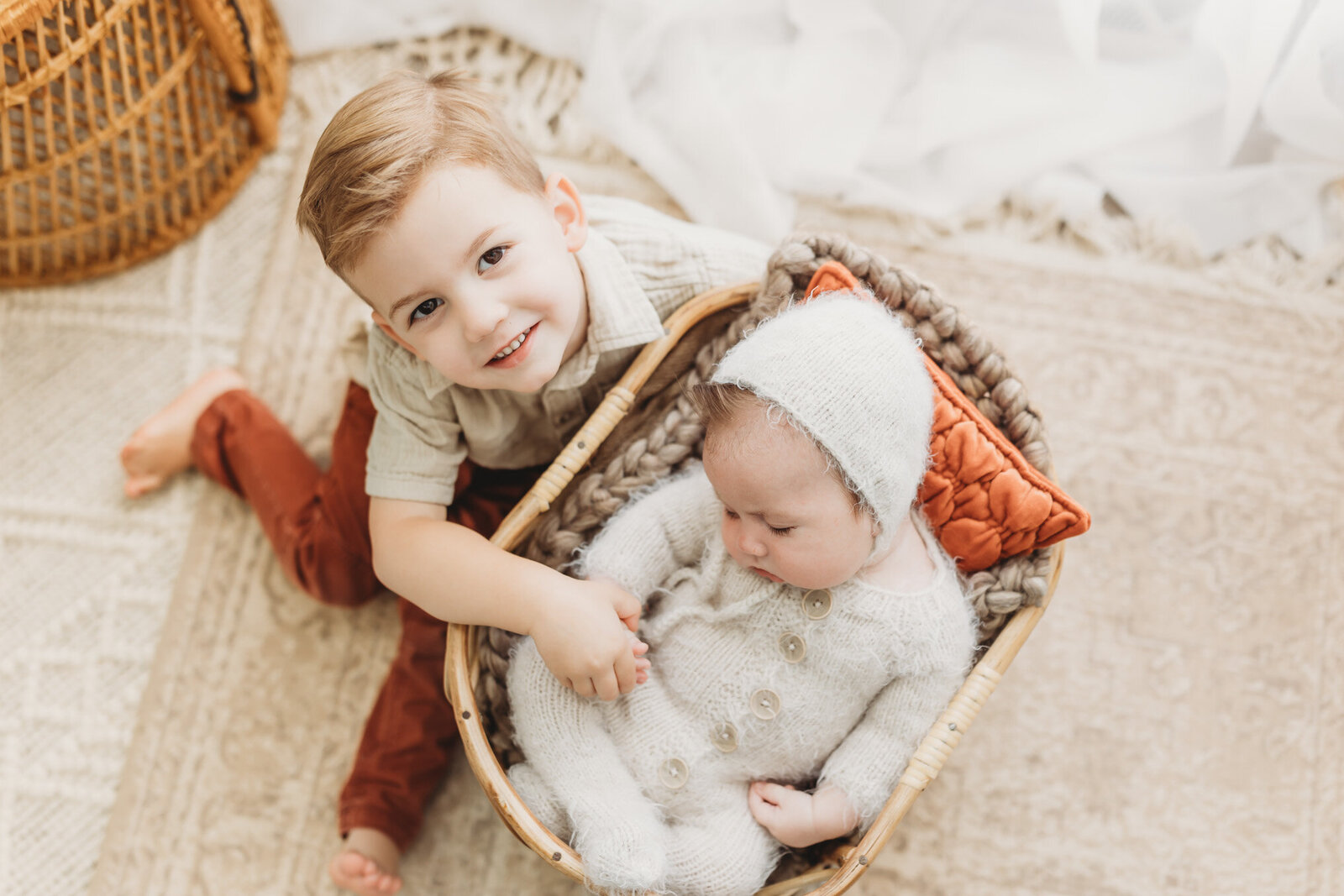 Toddler boy sitting next to newborn baby brother who is sleeping in a basket wearing a fuzzy bonnet and sleeper