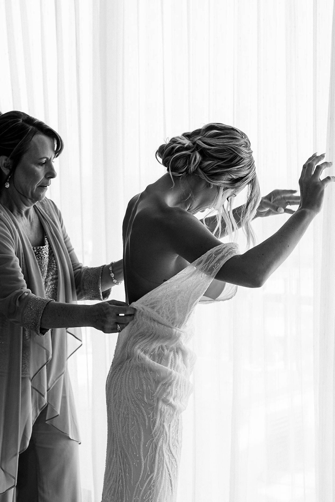 A beautiful black-and-white moment of a mother helping her daughter into her wedding dress as soft light filters through the window, captured by CMH Studios in San Antonio