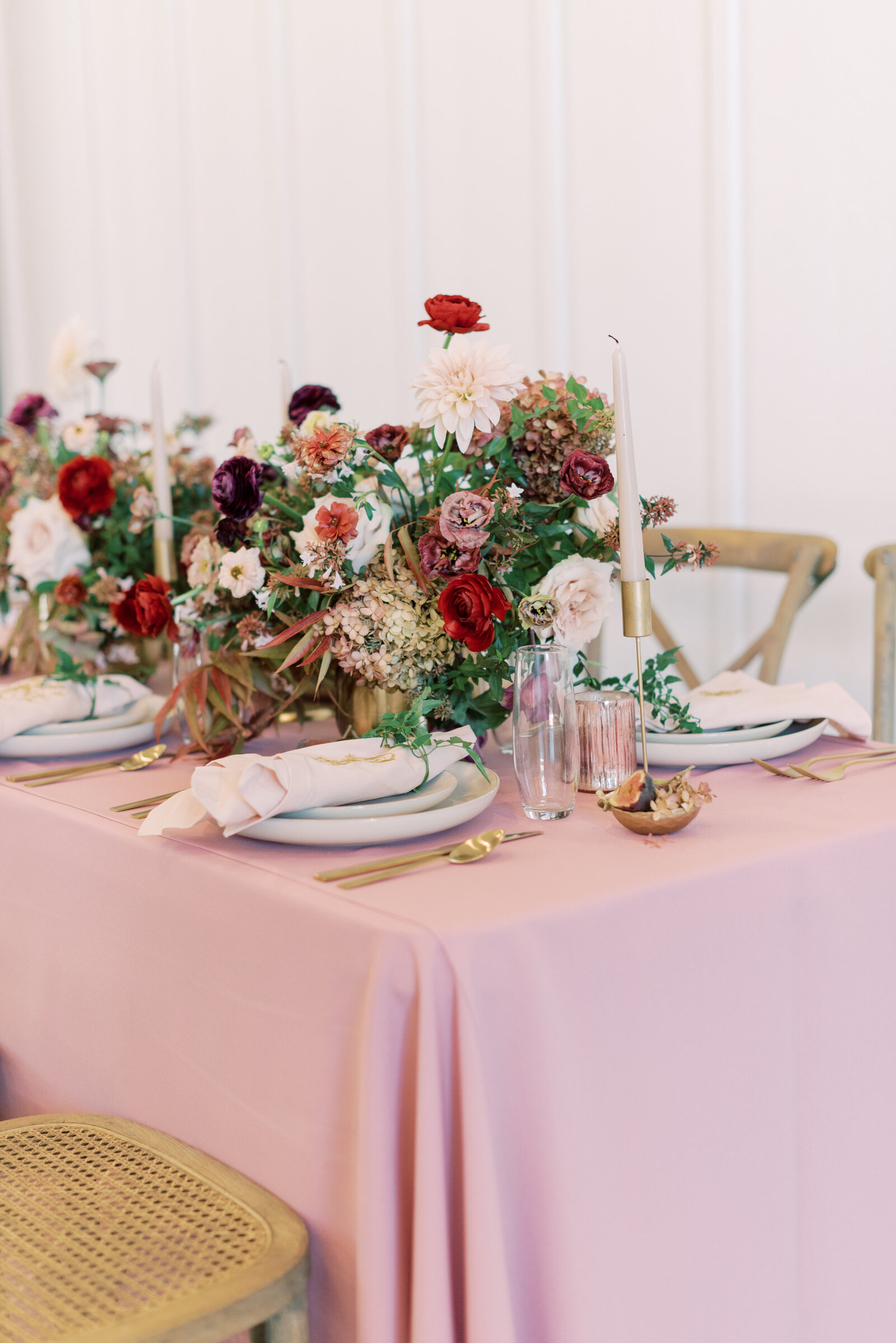 Table with gold silverware and ivory plates atop a table with pink linens, flowers, candlestick, and rattan chairs.