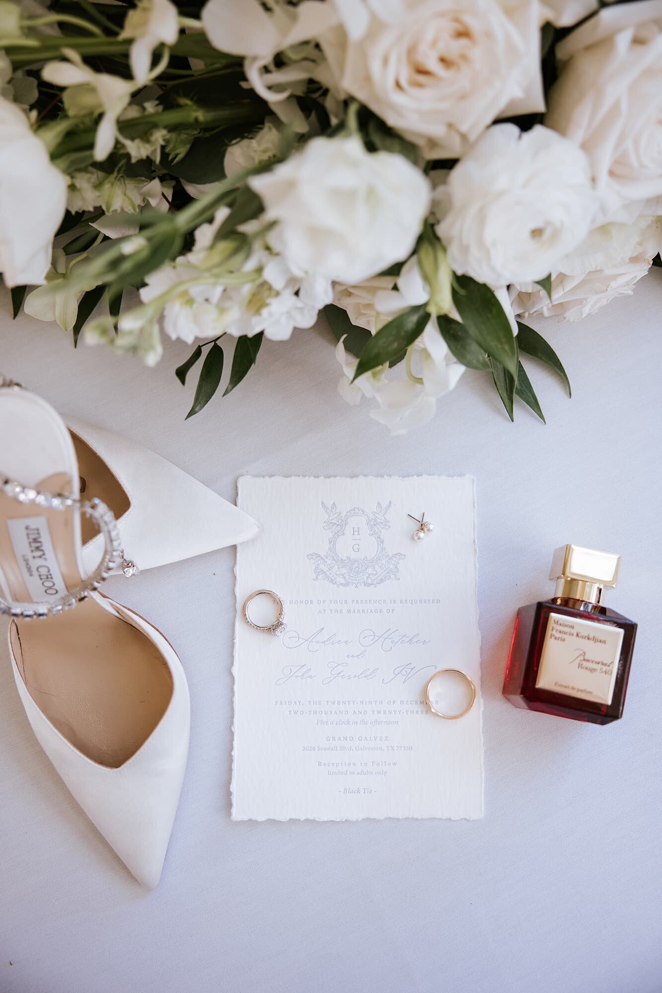 Luxury wedding flat lay featuring a white floral bouquet, Jimmy Choo bridal heels, gold wedding rings, a wedding invitation for a Grand Galvez ceremony, and a bottle of Maison Francis Kurkdjian perfume, styled by Houston wedding photographer Logrono & Co.