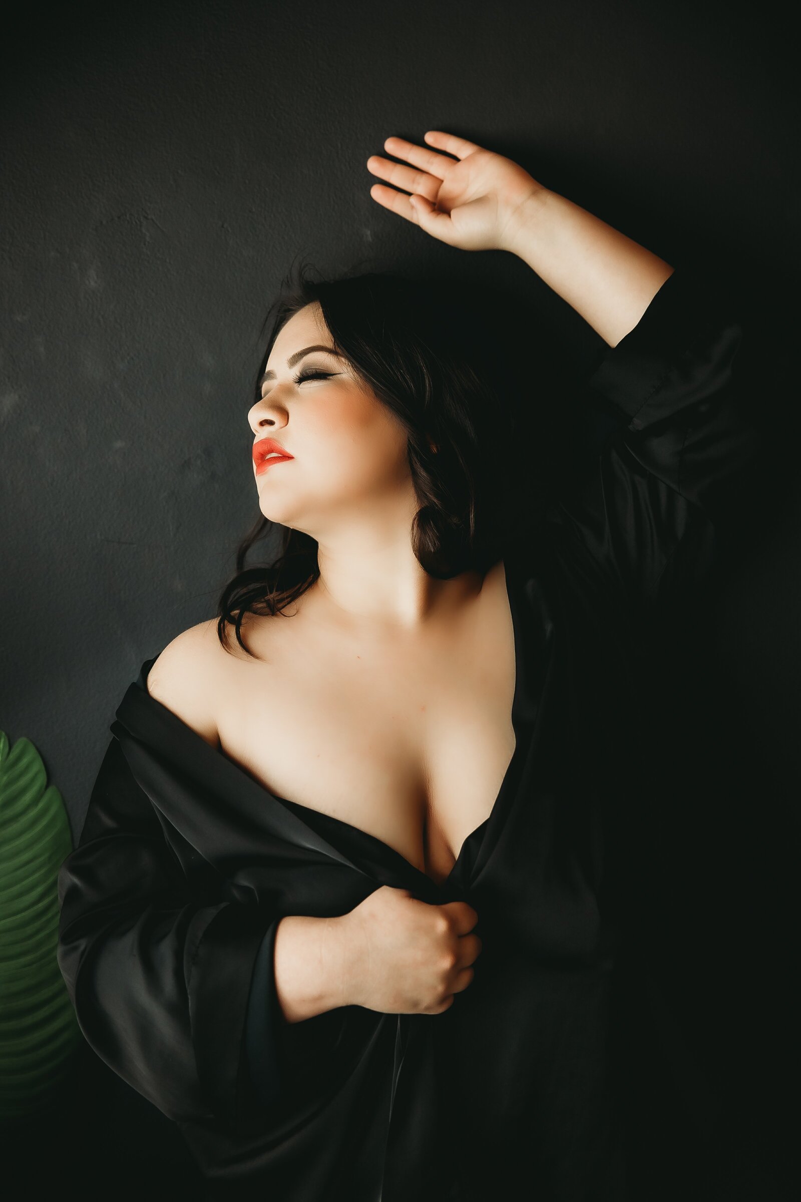 Travel location portrait of women in black silk shirt leans against black wall with her eyes closed and her left hand above her head