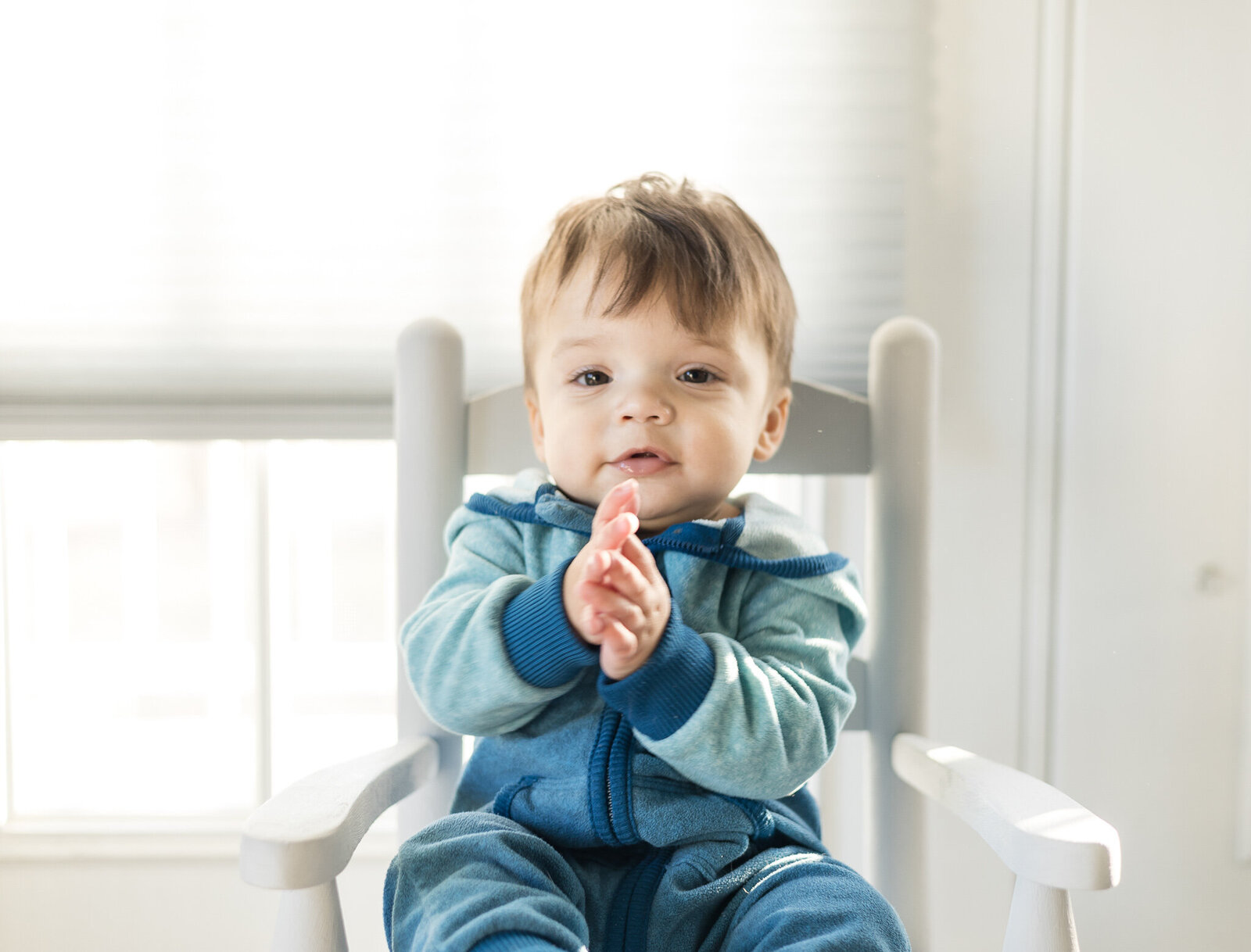 A 1 year old boy with dark brown hair sitting in a light gray rocking chair, with his hands together, looking at the camera, wear a blue zip up outfit