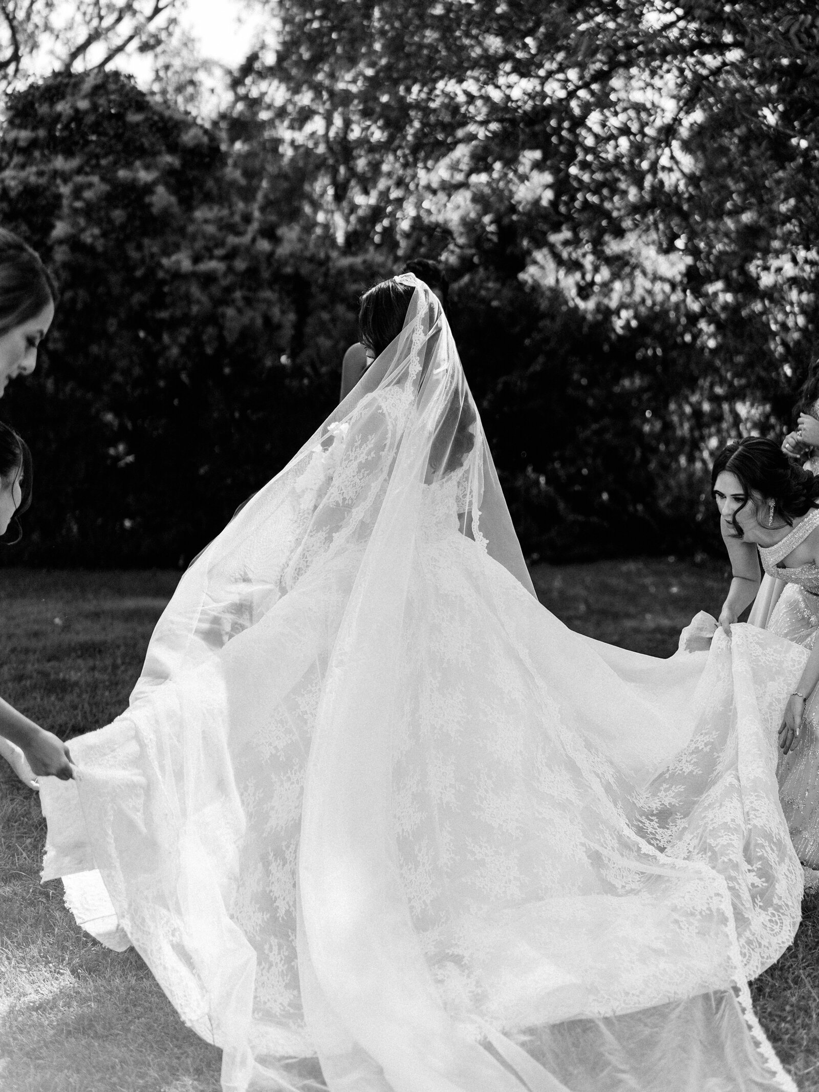 Black and white portrait of Bride surrounded by her bridesmaids  at Tudor Garden  as they adjust her veil and the wind sweeps it away.