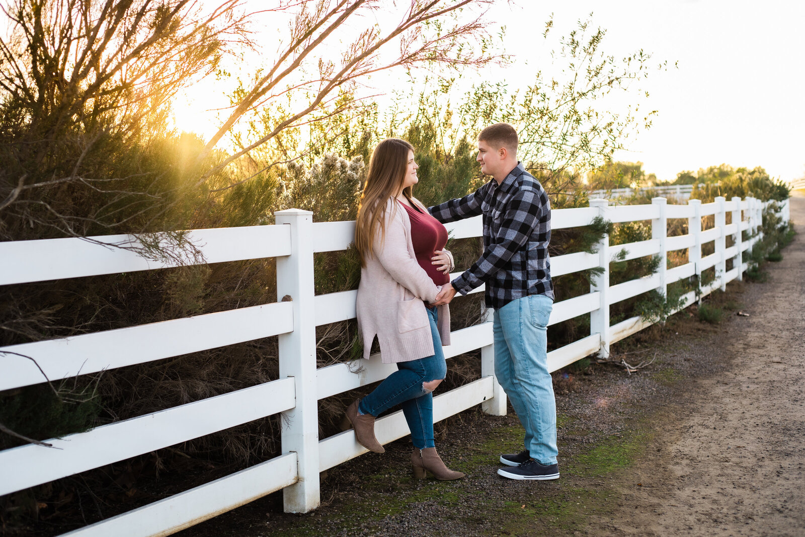 Maternity Photographer, a pregnant woman leans on a country fence, he holds her hands