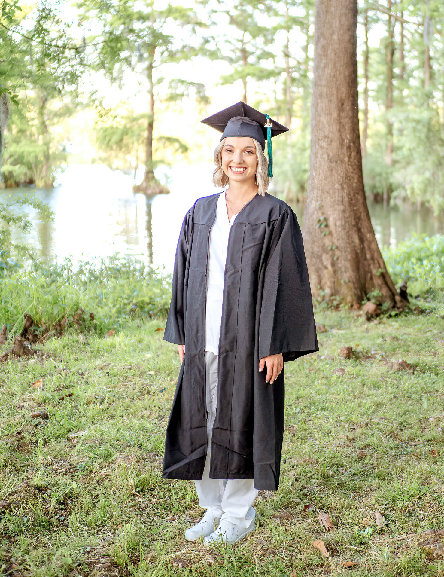 A smiling University of North Carolina Wilmington senior in a cap and gown standing in a lush lakeside setting in North Carolina. This photo captures the joy of graduation, perfect for those seeking beautiful outdoor senior photography in Wilmington.