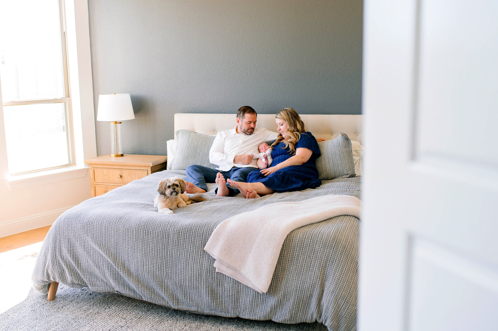 Father sitting on bed holding baby during a relaxed at-home newborn photography session in Prosper, Texas, captured by Jennifer L. Kirk Photography.