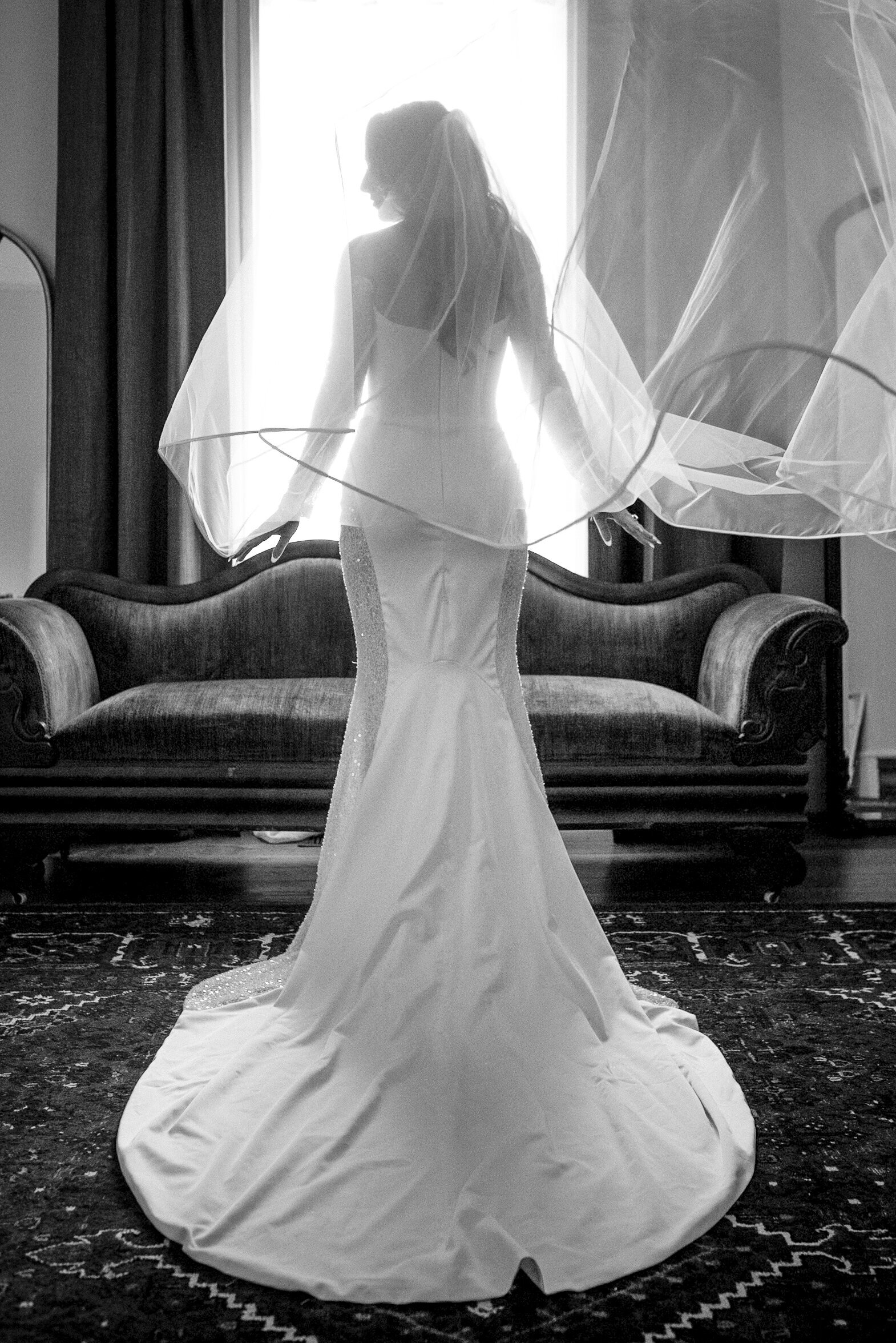 Black and white bridal portrait silhouette where the bride is facing away from the camera, standing in front of a window looking over her left shoulder, her veil floating in the air behind her. Photograph taken at The White Home in Rock Hill, SC.