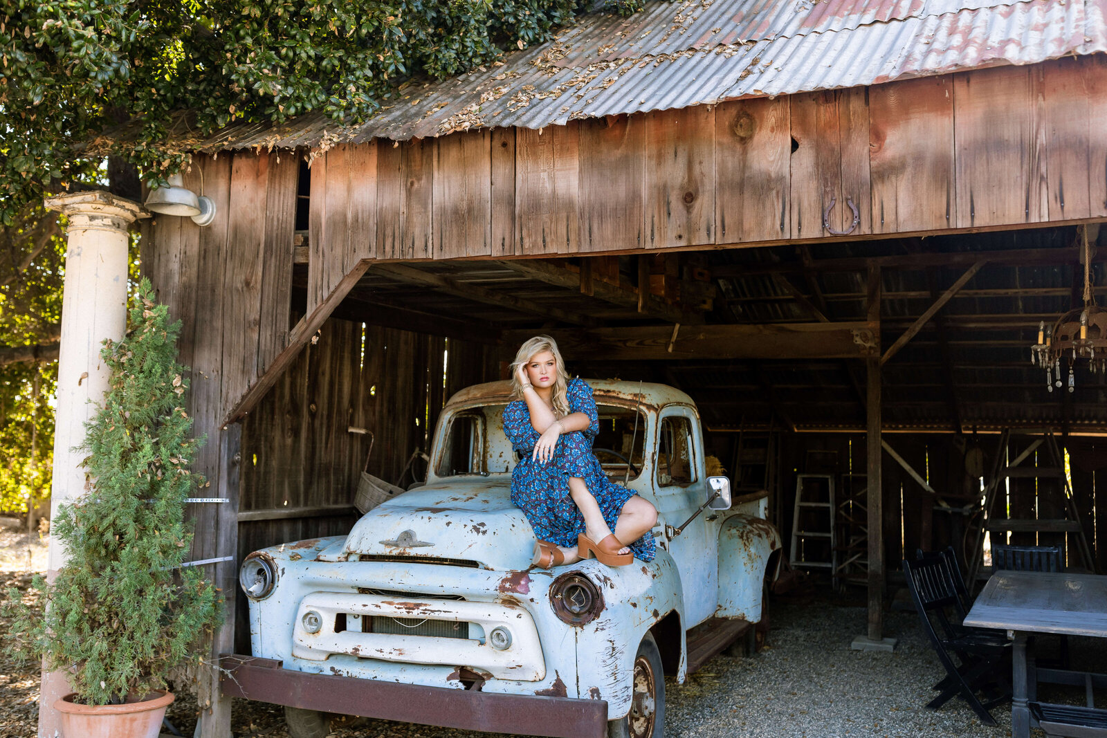blonde high school girl wearing a blue dress with brown heels posing on a blue rusted car under a barn