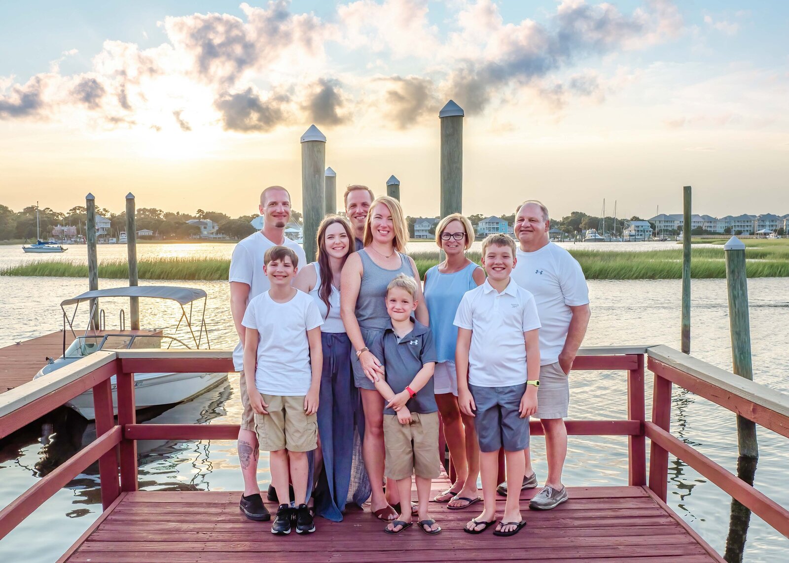 A large family poses together on a marina dock at sunset, dressed in white and khaki outfits. The boats and marshland provide a scenic coastal setting, perfect for professional family photography in NC, showcasing vibrant family gatherings.
