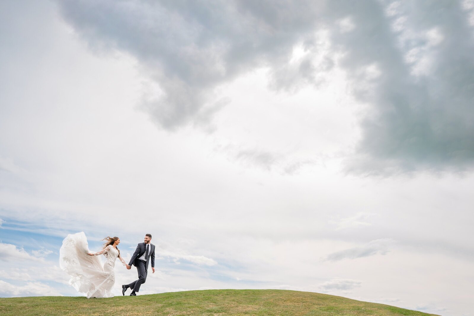 Bride and room running across a grassy hill with the sky and clouds in the background at Sleepy Ridge.