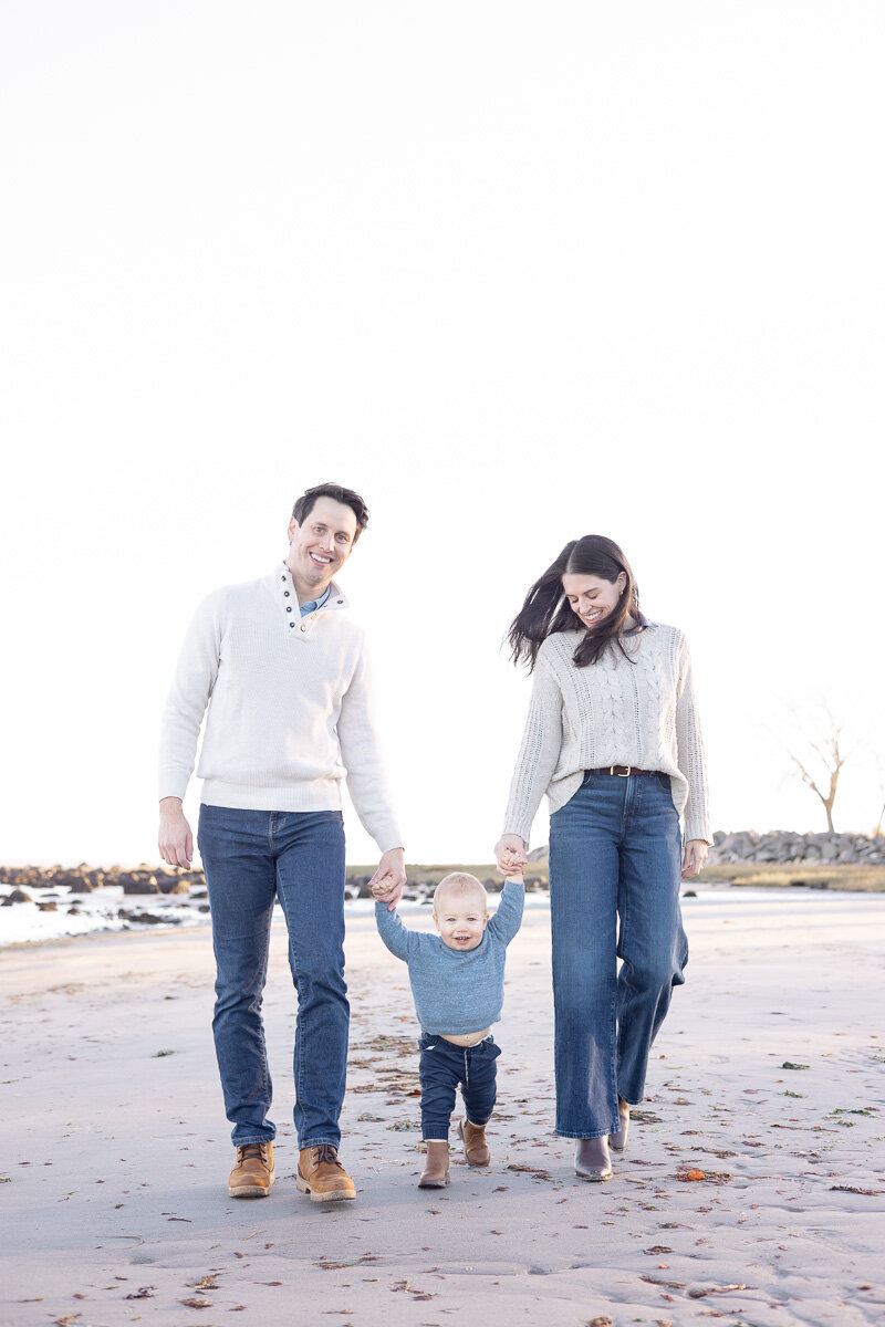 Young family holding hands and walking along the beach together in Westchester County, NY during their lifestyle family photography session. Captured by Nicole, a Fairfield and Westchester-based photographer for playful, connection-filled family moments.