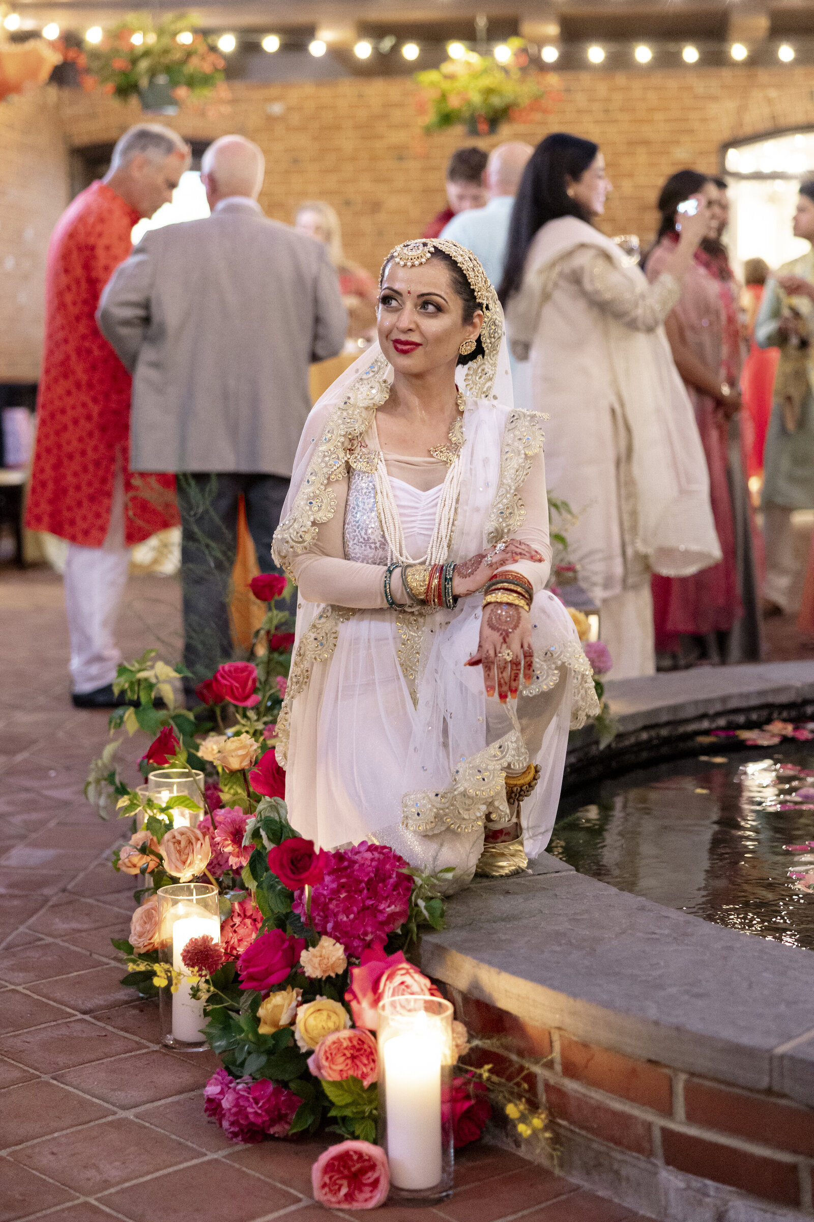 Woman sitting on the side of the fountain with wedding guests in the background and florals and candles to her side.
