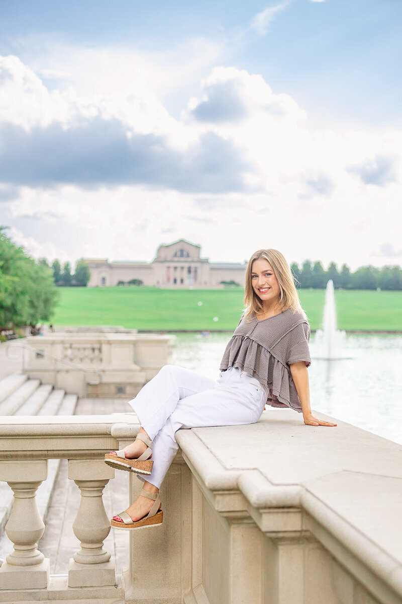 senior sitting on railing at grand basin in forest park with st. louis art museum in background
