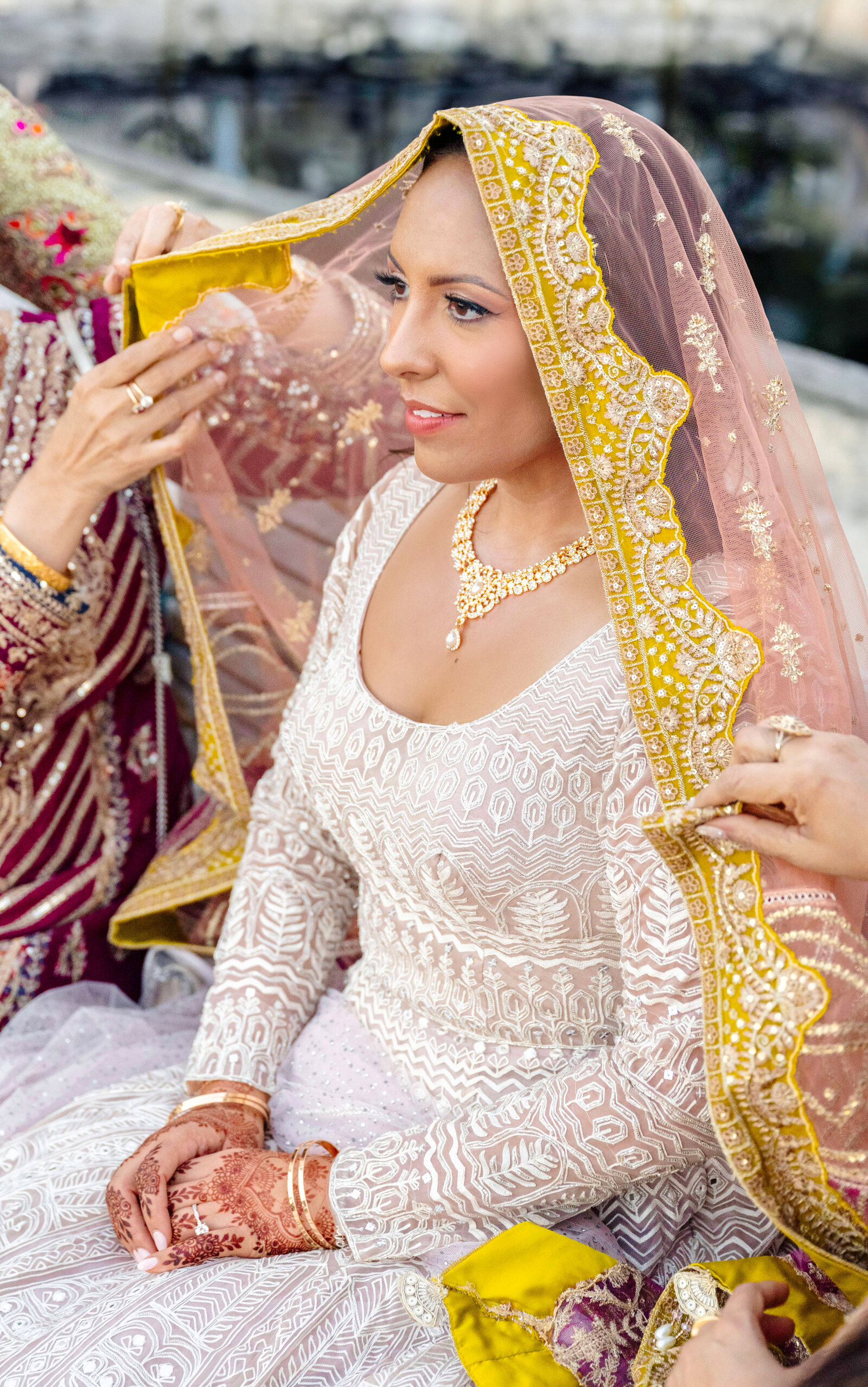 A Pakistani bride in a detailed blush dress is having a golden and pink lace veil placed on her head by two women. She wears intricate jewelry and henna on her hands, seated in an ornate setting.