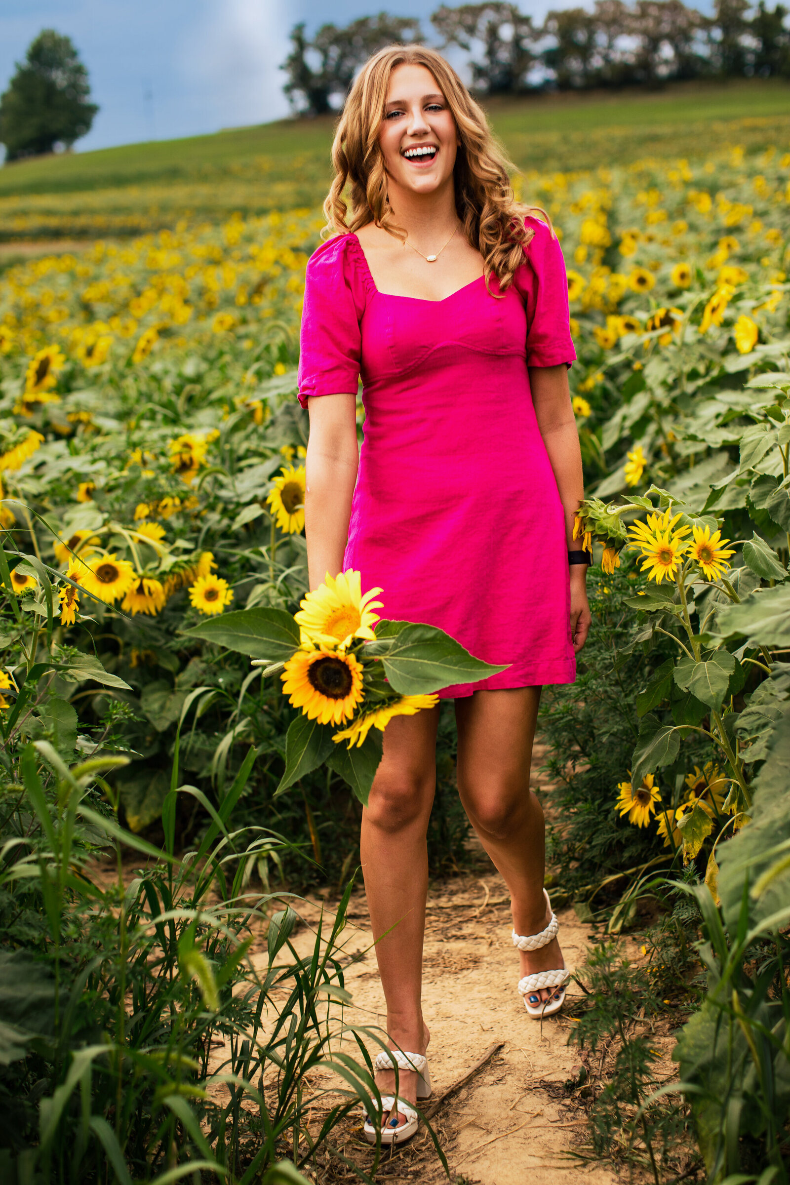 A girl wearing a bright pink dress and beautiful White sandals walks through a sunflower field holding a bouquet of sunflowers in her hand.