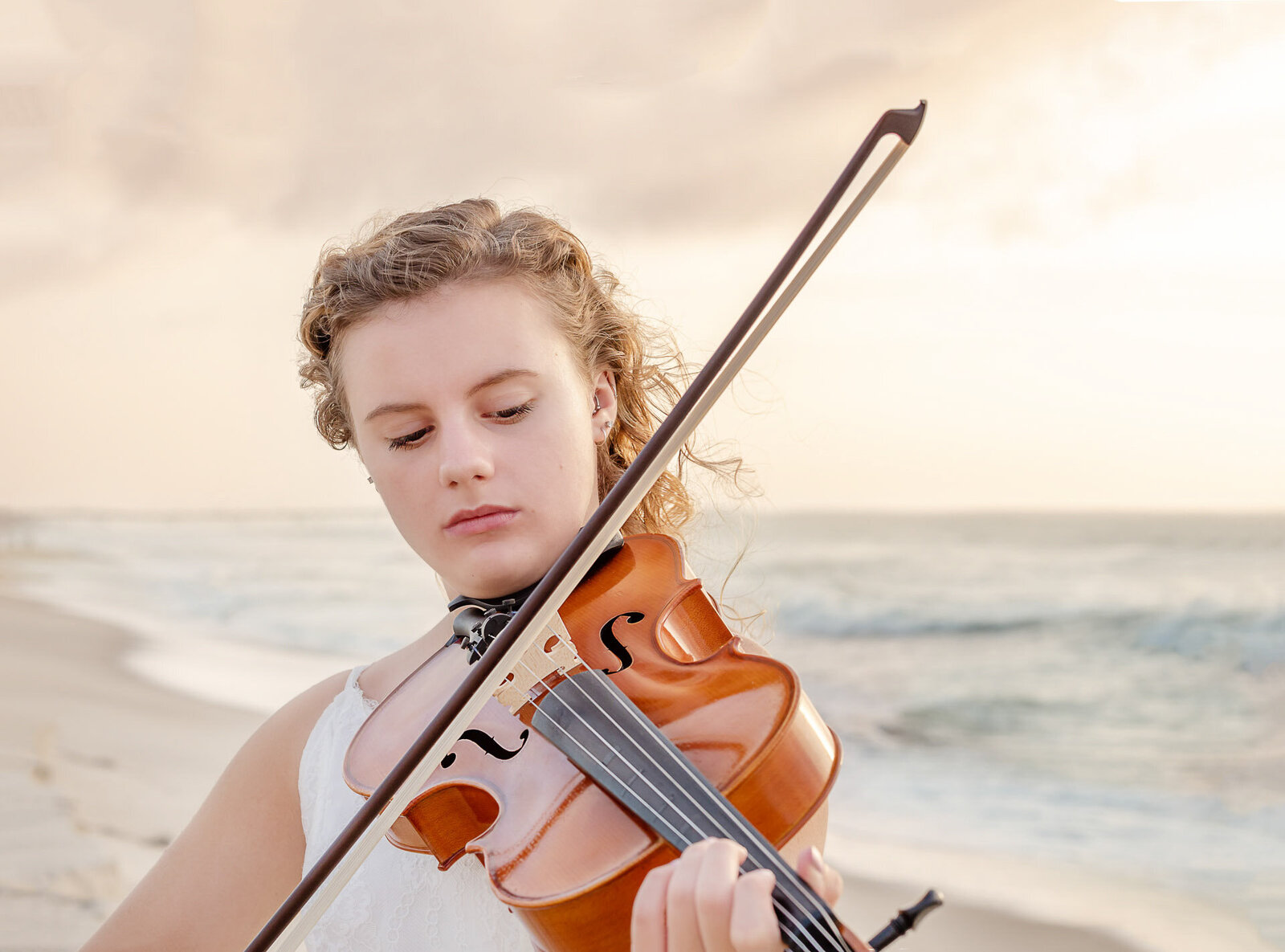 A high school senior in a white dress playing the violin on serene Carolina Beach in North Carolina. This artistic portrait captures the harmony of music and nature, ideal for those seeking unique senior photography near Wilmington, NC.