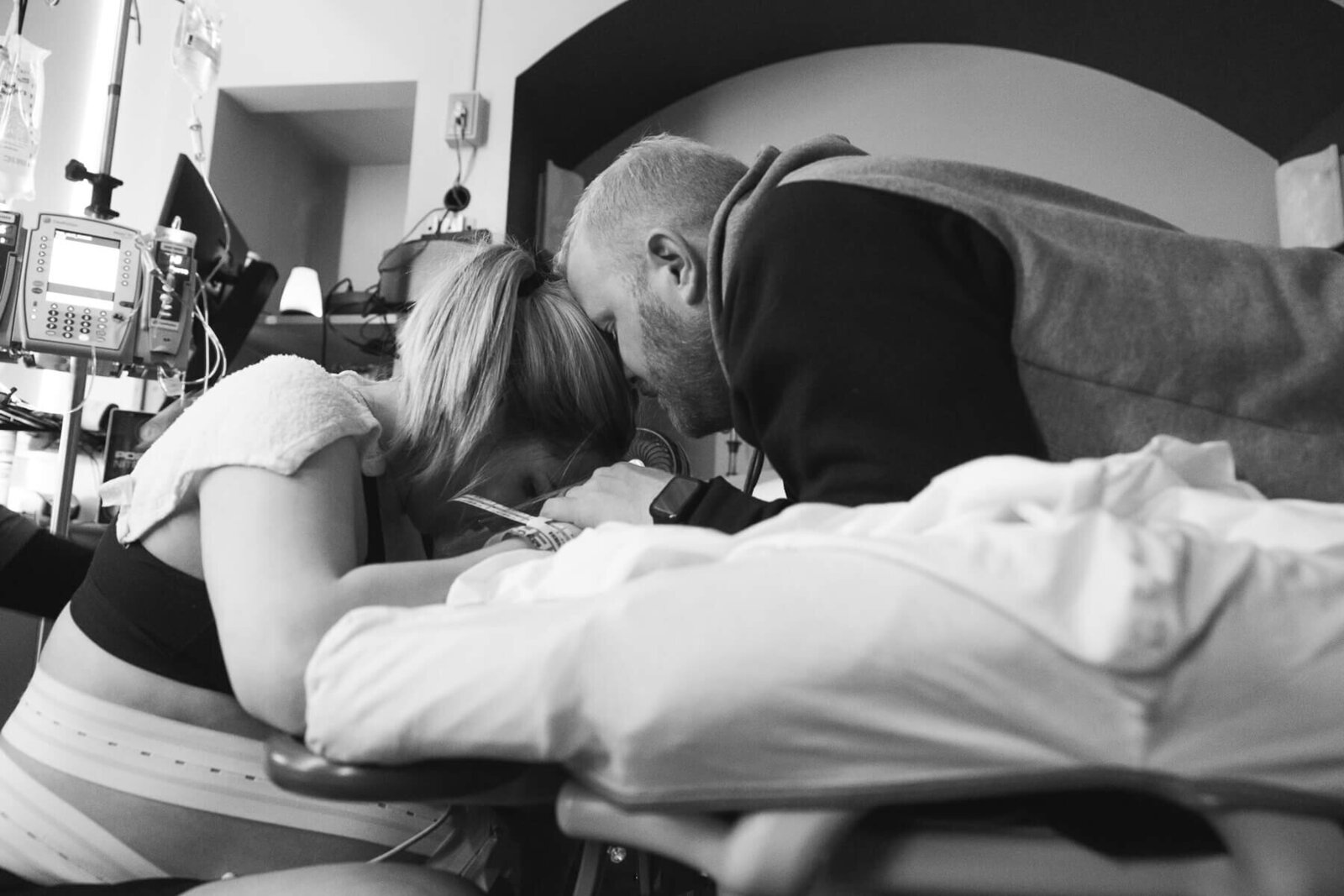 A man leaning over a hospital bed and leaning his head against his wife's head comforting her as she is in labor in the hospital.