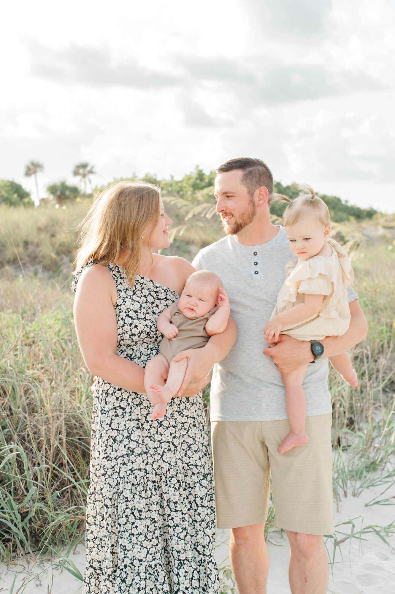 Family of four in beautiful green tones stands near the dunes smiling at each other during their time with a Cocoa beach family photographer.