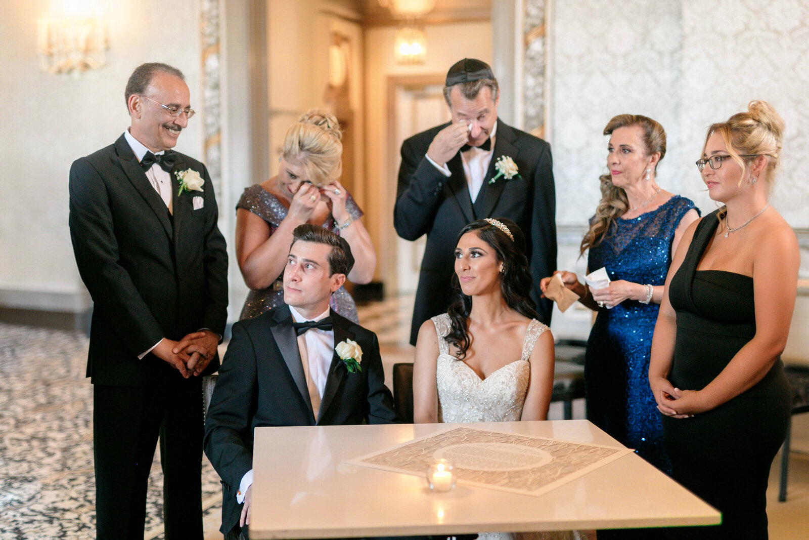 A Persian bride and Jewish groom sit at a table with a candle, surrounded by family members. The group, dressed formally, appears emotional inside an elegantly decorated room with ornate details and chandeliers.