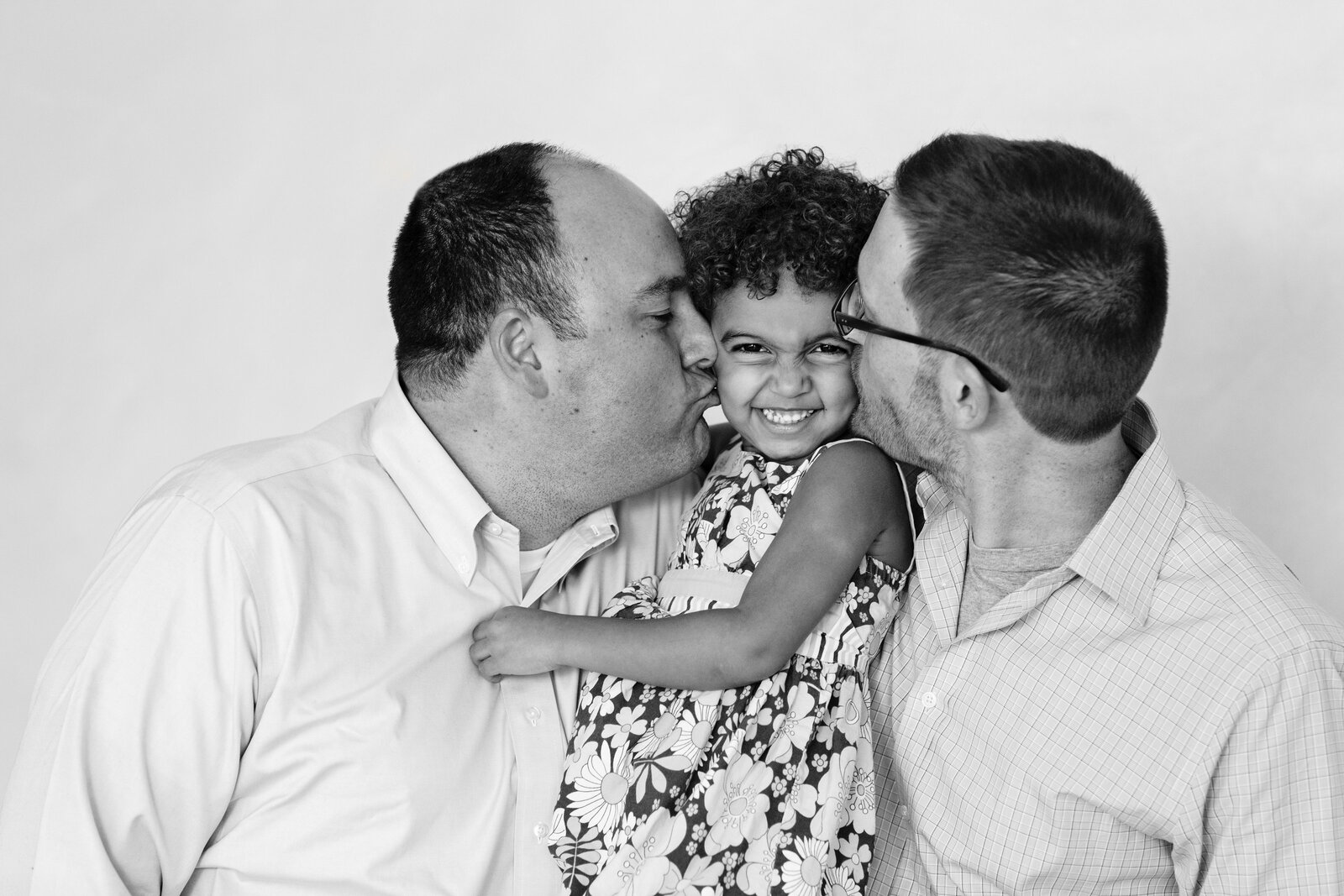 Family of three posing together, with gay dads hugging their smiling daughter who looks at the camera giggling