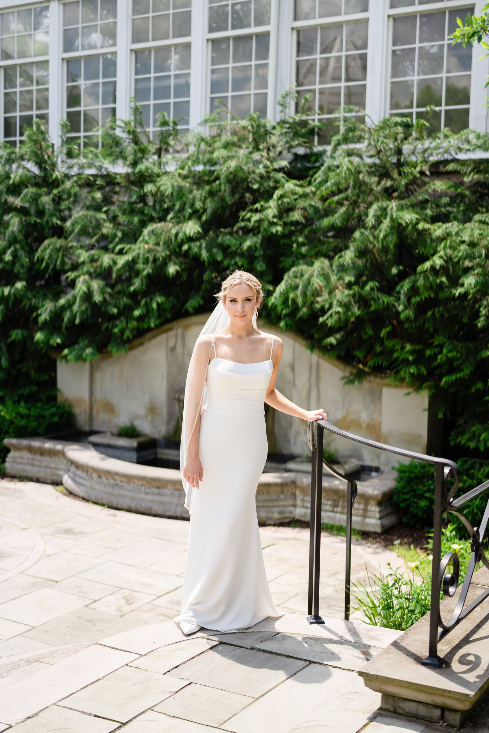 A bride in a white dress holds onto the rail and softly smiles in the Bride's Garden at the Franklin Park Conservatory