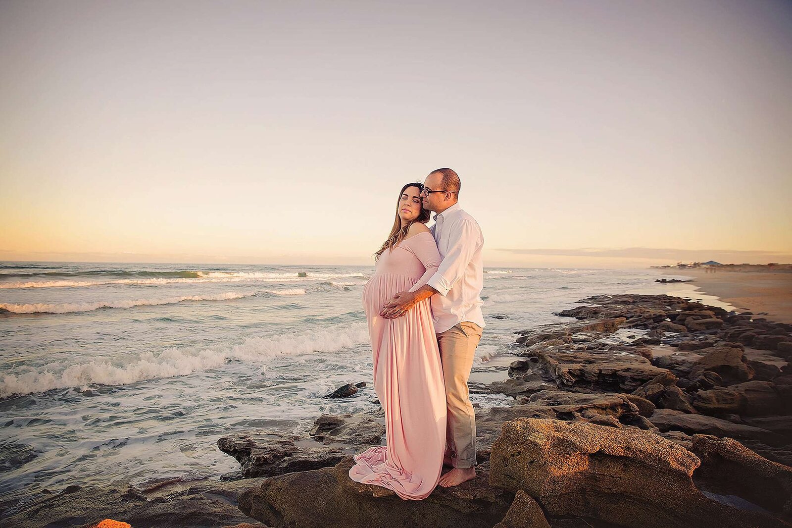 A pregnant couple stands on rocky beach terrain, capturing a serene moment during their maternity session by Bia Schaefer Photography.
