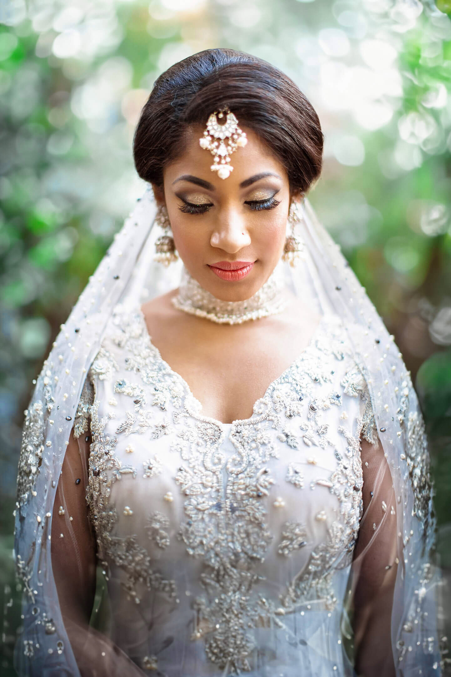 An Indian bride in an ornate light blue gown with intricate beadwork and lace stands outdoors. She wears a traditional headpiece, earrings, and a necklace. Her eyes are closed, and her veil drapes gracefully over her shoulders, framed by soft, blurred greenery.