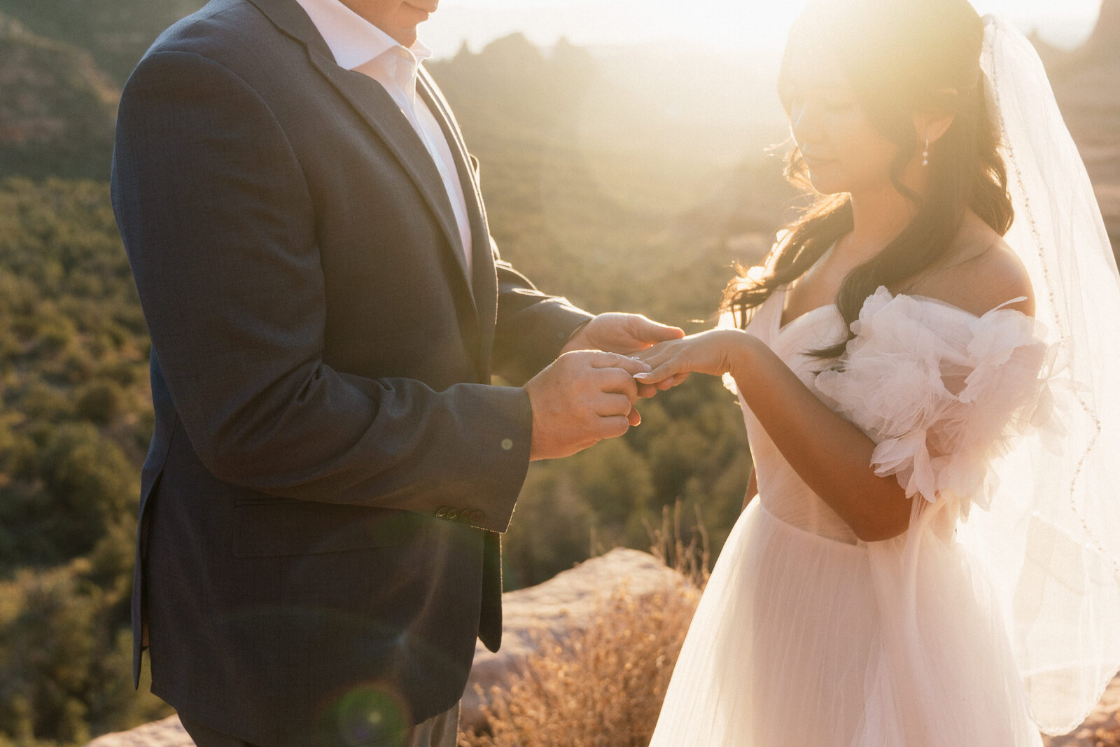 Groom placing a ring on the bride's finger during a golden hour elopement ceremony at Merry Go Round Rock in Sedona, Arizona, with warm sunlight illuminating the desert landscape in the background.